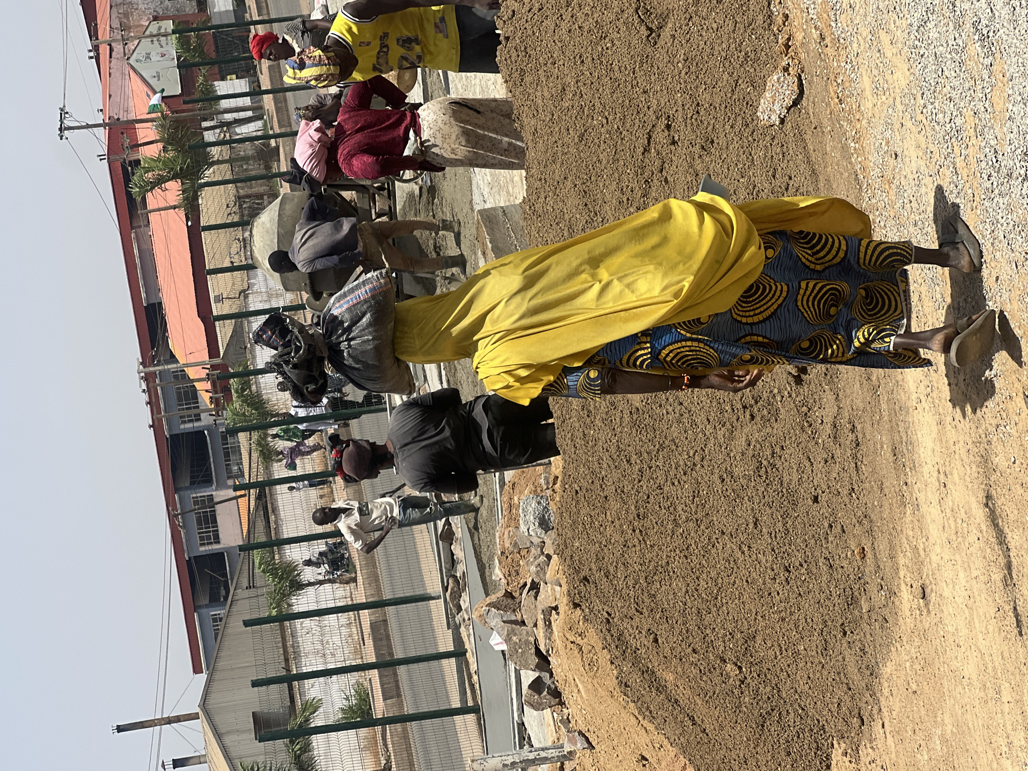 In Barakin Lerawi, Nigeria, the photograph depicts a bustling construction site adjacent to a gated perimeter surrounding a building. A woman in a yellow garment and a blue and yellow patterned skirt stands near a mound of brown earth, carrying a heavy sack atop her head. Several other people are visible, some appearing to operate a cement mixer. The background includes buildings and utility poles, suggesting an urban environment. The ground is a mixture of sand and gravel.