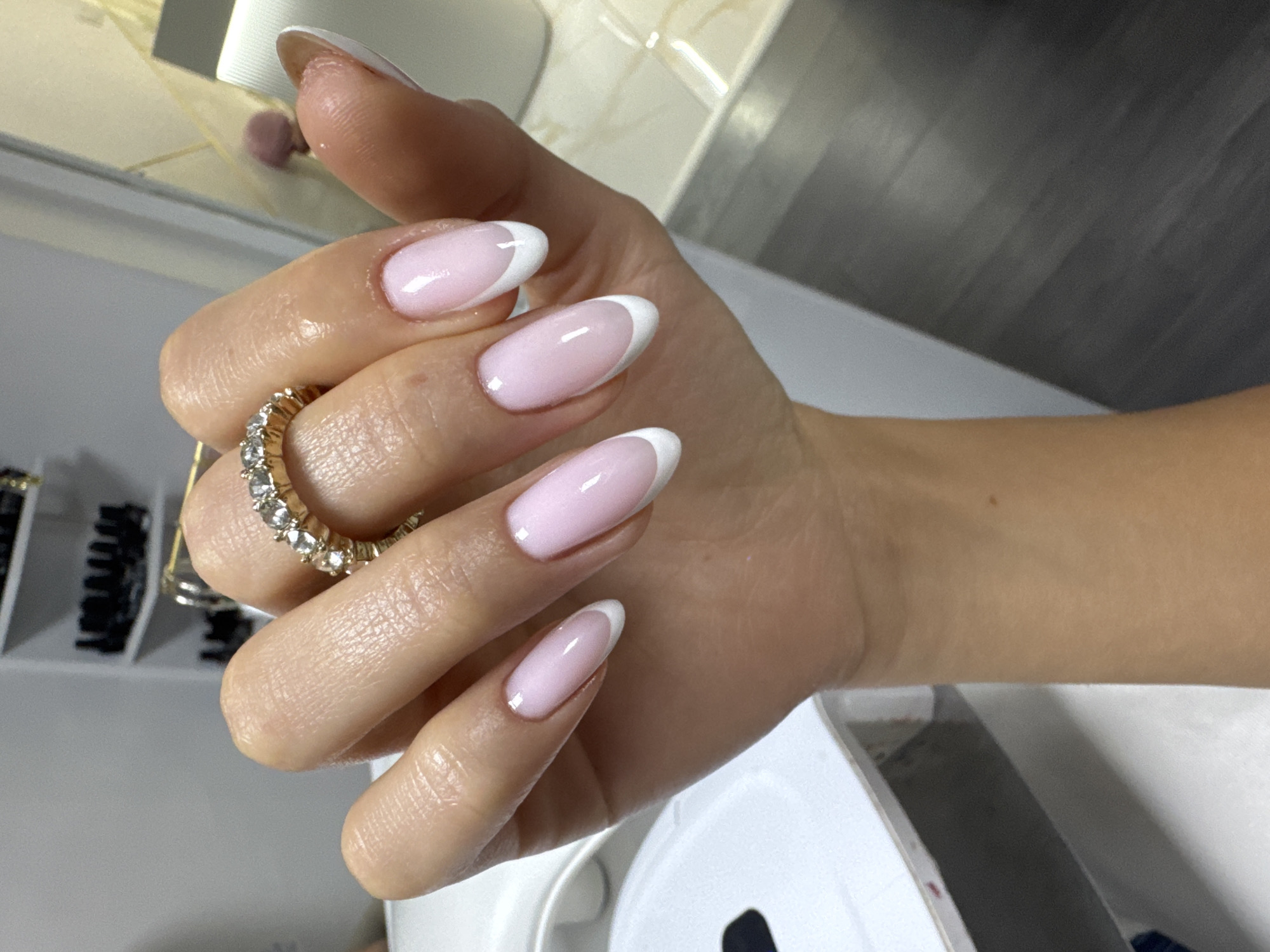 A close-up photograph shows a woman's hand with manicured nails featuring a classic French tip design (pink base, white tips), adorned with a gold-colored ring set with clear stones. The hand is positioned against a background that includes a portion of what appears to be a white or light-colored appliance (possibly a nail lamp) and a blurred shelf or container holding hair accessories. The location cannot be determined without additional contextual clues.