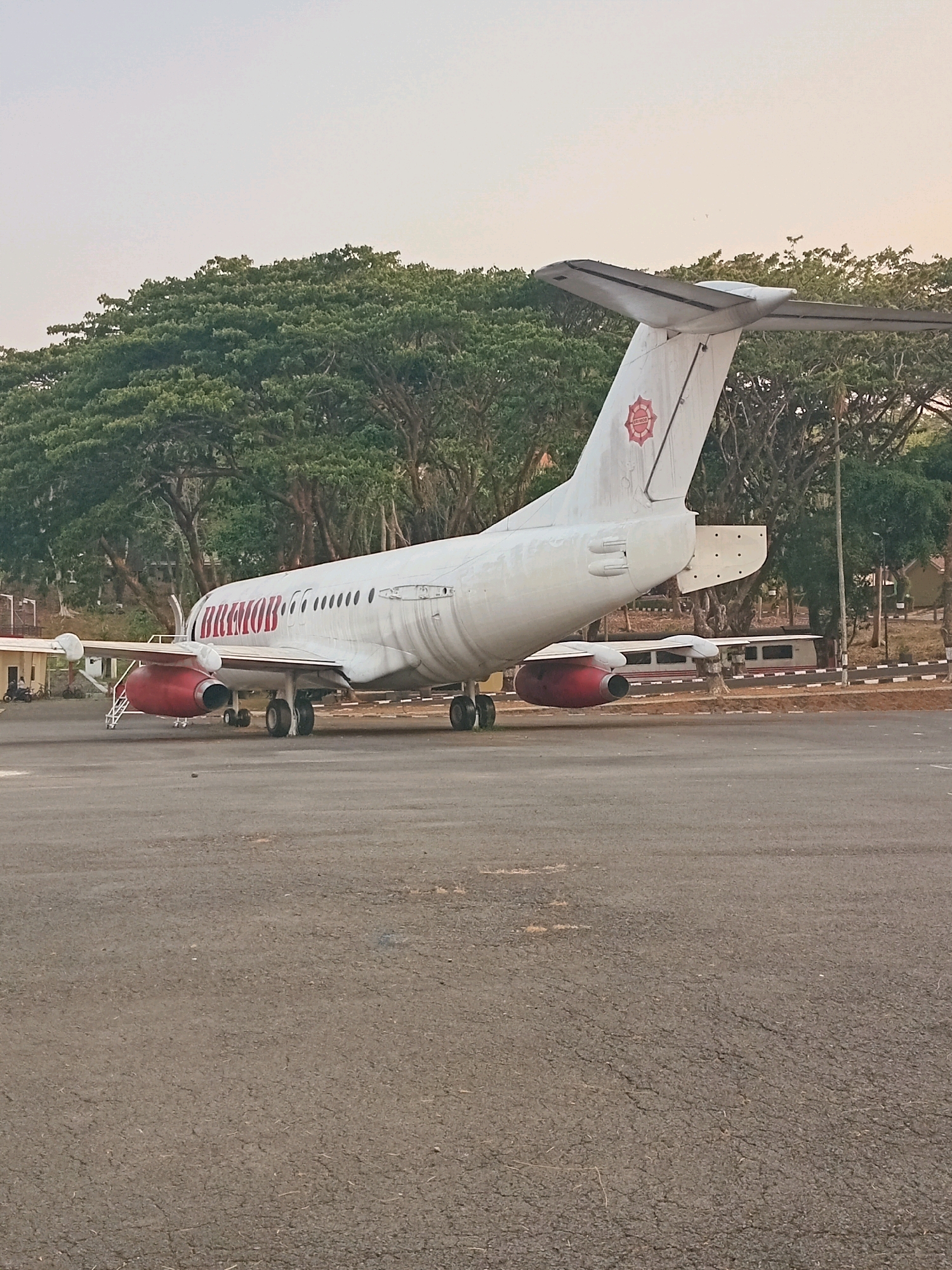 A stationary white airplane with red accents is parked on an asphalt tarmac. The airplane has "BRIMO" written in red letters along its fuselage. Its engines are red, and it has a twin-engine configuration. The tail of the plane features a red circular insignia with a star-like design. In the background, a line of lush green trees forms a dense canopy. Beyond the trees, some buildings are partially visible. The sky is a pale, overcast gray. The scene is devoid of any visible human activity or interaction. The image is taken from a low angle, looking towards the airplane. The location is not identifiable as Semarang, Indonesia based on the visual information provided.