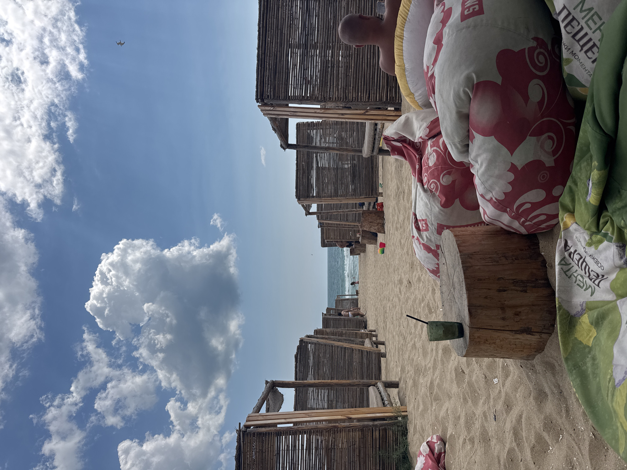 The image depicts a serene beach scene in Nesebar, Bulgaria, characterized by several wooden beach cabanas with thatched roofs lining a sandy shore. In the foreground, several large beanbag chairs, some with floral red and white patterns and others with green, are arranged on the sand. A wooden log serves as a table, with a glass containing a dark liquid and a straw resting on it. The sea is visible in the background, with gentle waves and a hazy horizon. The sky is blue with scattered white clouds, and a bird is seen flying in the distance. A few people are present on the beach, some near the cabanas and others further down by the water. The overall atmosphere is one of relaxation and leisure on a sunny day.