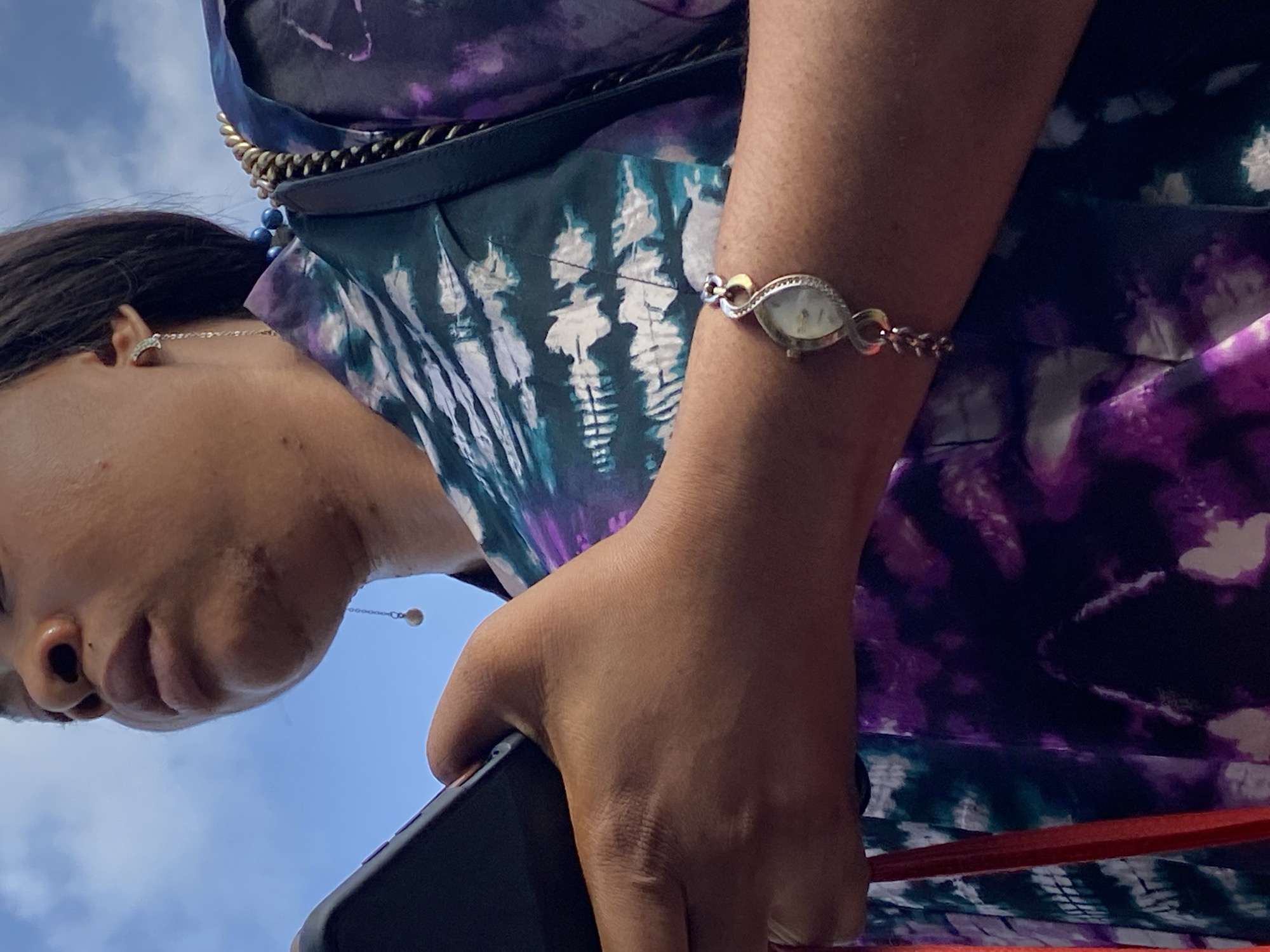 A person stands outside under a blue sky with some clouds, likely in Lagos, Nigeria based on the skin tone of the person and the sky's hue. The individual is holding a black smartphone in their right hand. They are wearing a purple, white, and green patterned dress with a gold chain strap. A decorative silver watch adorns the person's right wrist. They wear stud earrings and a dangling earring on the left side.