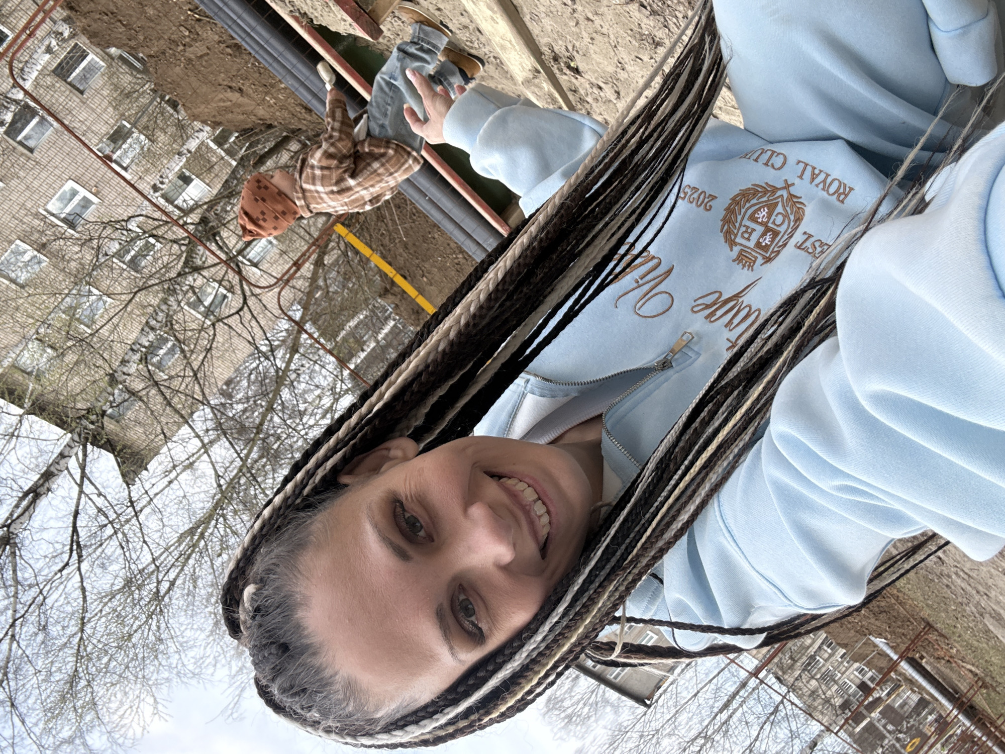 The image features a selfie-style shot of a smiling person looking directly at the camera. They have long, multi-toned braided hair, including dark brown, black, and lighter blonde/grey sections. The person is wearing a light blue zippered hoodie with prominent embroidery. Visible text on the hoodie includes "ROYAL CLUB", "EST. 2025", a crest logo, and a larger cursive word that appears to be "Vintage" or similar, beginning with a 'V'.
In the background, another person, possibly a child, is partially visible. This individual is wearing a brown plaid shirt, a brown knitted hat, and blue jeans, and appears to be sitting or playing on a low structure made of metal pipes and wooden components on a dirt ground.
The scene is outdoors in what seems to be a residential or urban environment in Rusanovo, Russia. A large, multi-story building made of light-colored bricks or concrete, featuring many windows, is visible in the distance. Bare tree branches are scattered throughout the frame, suggesting a colder season such as late autumn, winter, or early spring. The sky is overcast, providing soft, diffused lighting. The main subject's expression is happy and friendly. The image is rotated horizontally.