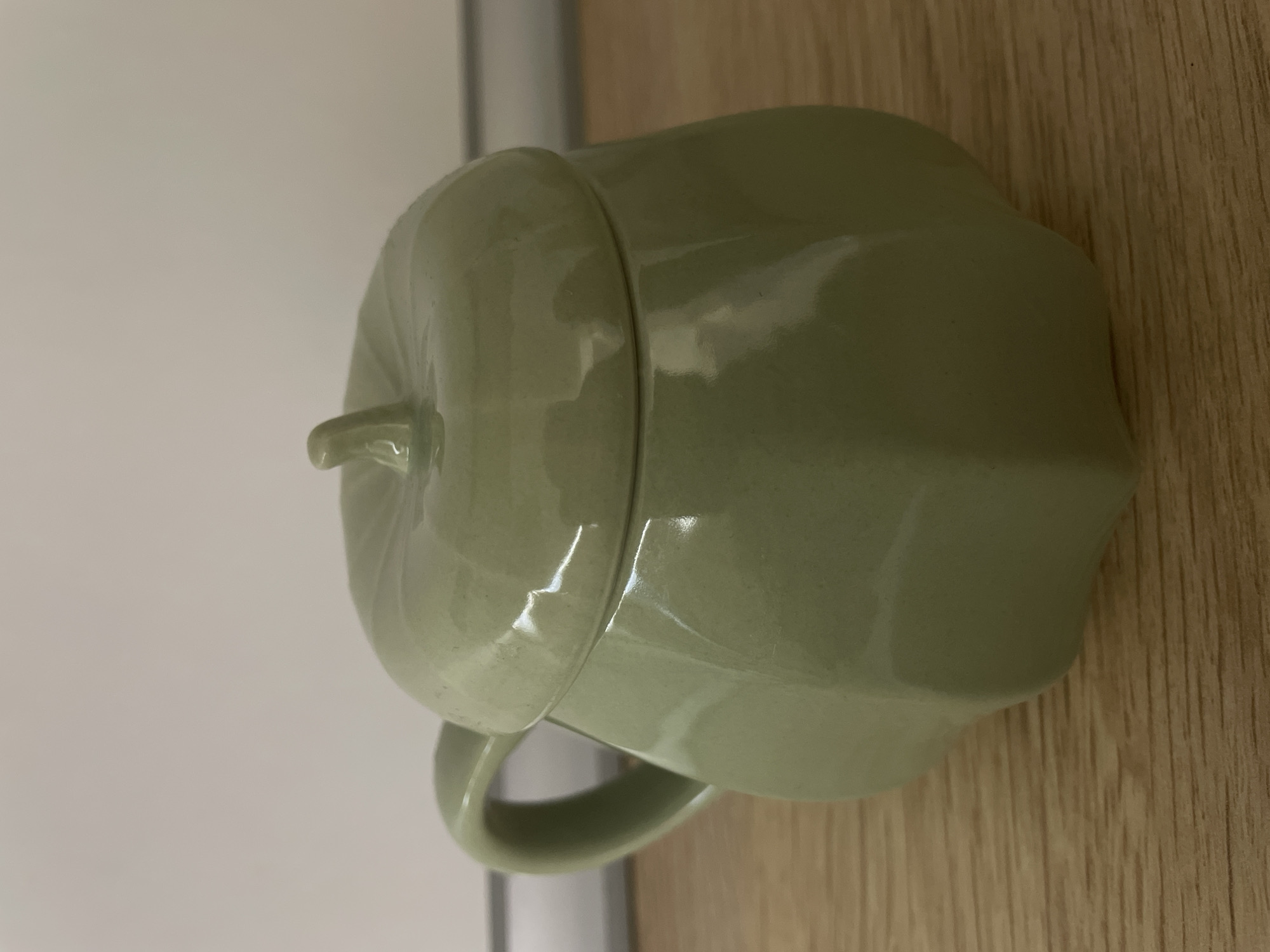 The image shows a close-up, eye-level, medium shot of a pale green ceramic mug shaped like a pumpkin. The mug has a lid with a small stem-like protrusion on top, and a handle is visible on the left side. The mug is placed on a wooden surface, which appears to be a table or shelf, with a plain, light-colored wall in the background. The lighting suggests it is indoors, possibly in a domestic setting. There are no people or any other objects in the frame. The image does not provide any information that could indicate its location in a city in Russia or any other geographical location.