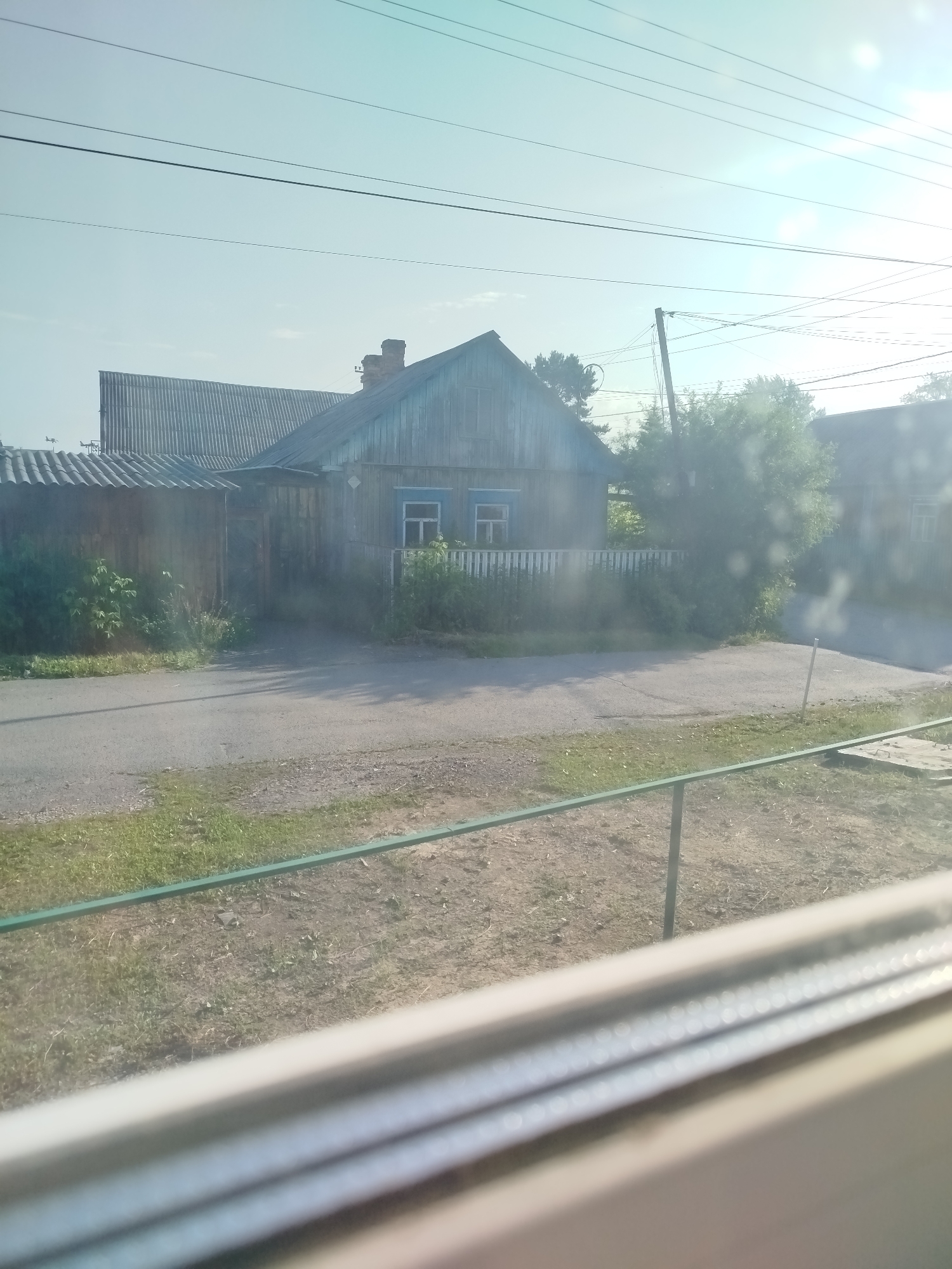 The image shows a weathered, wooden house with a corrugated metal roof, typical of rural Russian architecture, set against a bright, hazy sky. The house is partially obscured by overgrown vegetation and a wooden fence. A paved road runs in front of the property, with a grassy verge and a metal railing visible in the foreground. The overall scene suggests a quiet, perhaps slightly neglected, residential area. The presence of utility poles and wires indicates that the location is connected to infrastructure. The bright sunlight and haze suggest it might be a warm, summer day. The image is taken from a slightly elevated perspective, possibly from a window. The city of Zavodoukovsk is known for its industrial areas, but also has residential districts with similar housing styles.