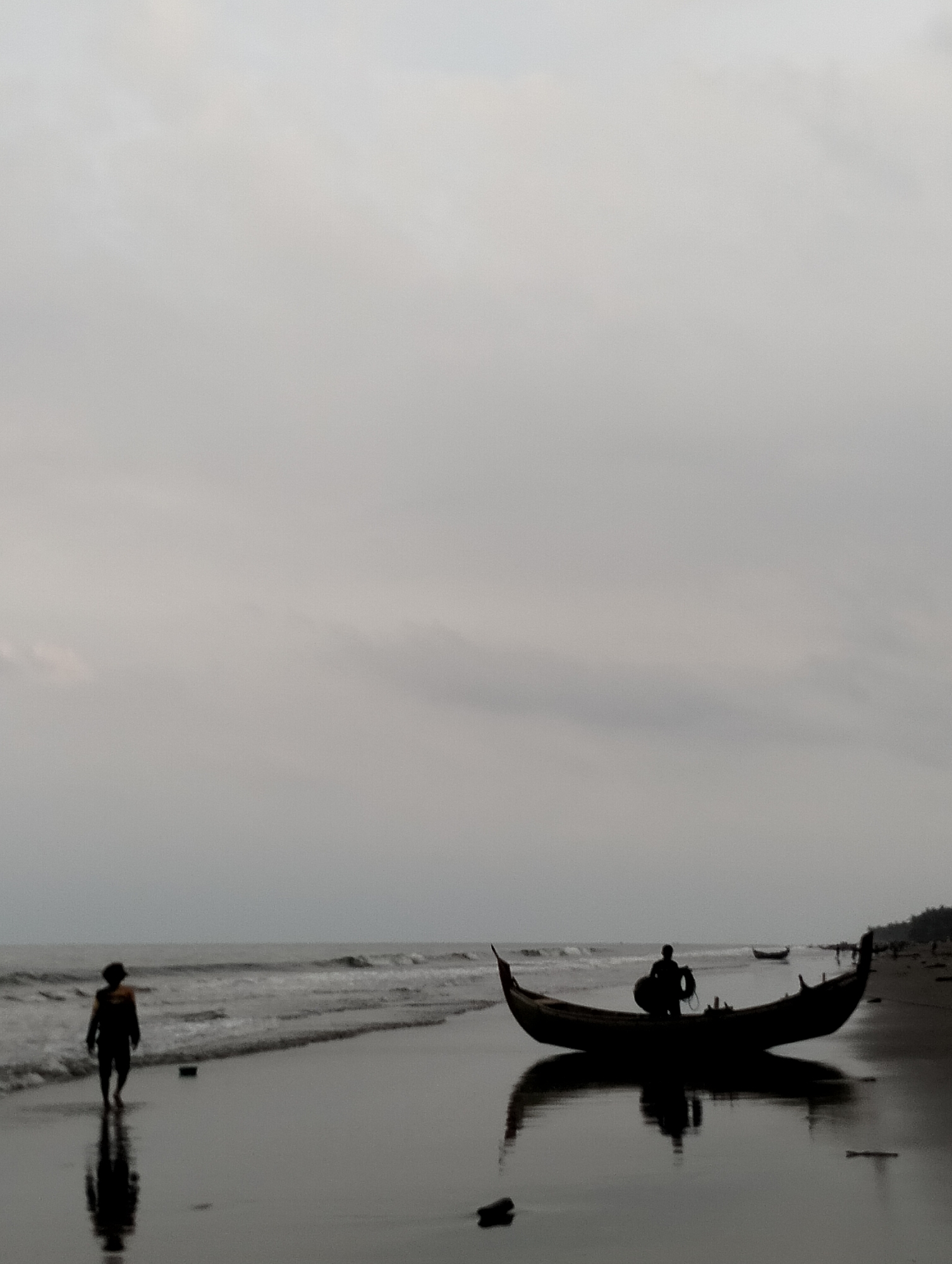 The image depicts a serene, overcast scene at a beach in Peuneulet Baroh, Indonesia. In the foreground, the wet sand reflects the dim light, creating a mirror-like surface. A lone figure, silhouetted against the lighter horizon, walks along the shoreline, their reflection stretching out before them. Further down the beach, a traditional Indonesian fishing boat rests on the sand, its silhouette also stark against the backdrop. A second figure is visible standing within this boat, holding what appears to be a life preserver. The ocean stretches out to the horizon, with gentle waves lapping at the shore. In the distance, a line of trees marks the edge of the land, and a few other indistinct figures can be seen along the coastline. The overall mood is quiet and contemplative, evoking a sense of solitude and connection with nature.