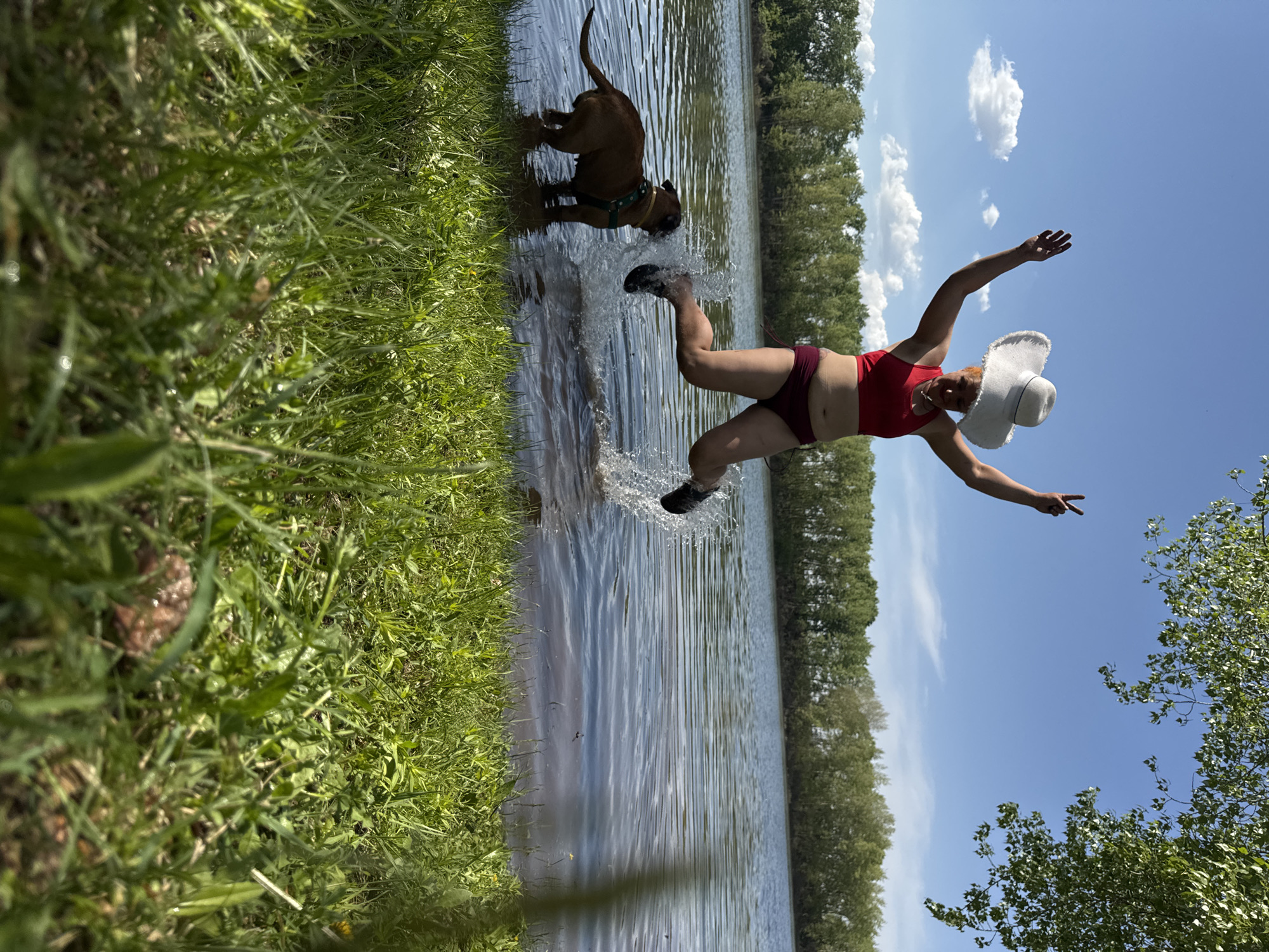 A woman wearing a red bikini and a white hat is captured in mid-air, leaping from the edge of a body of water. She holds one hand up with a peace sign. A dog is standing at the water's edge. The background shows a line of trees and a blue sky with scattered clouds. The location is Chaikovsky, Russia.