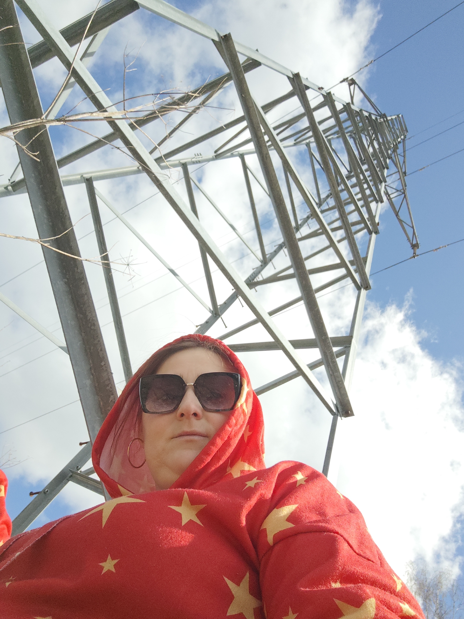 A low-angle shot captures a woman in a red, star-patterned hoodie and sunglasses, standing beneath a metal electricity pylon. The pylon towers above, intersecting with a partly cloudy, blue sky. Bare tree branches are visible, adding to the natural elements present. The location is Chaikovsky, Russia.