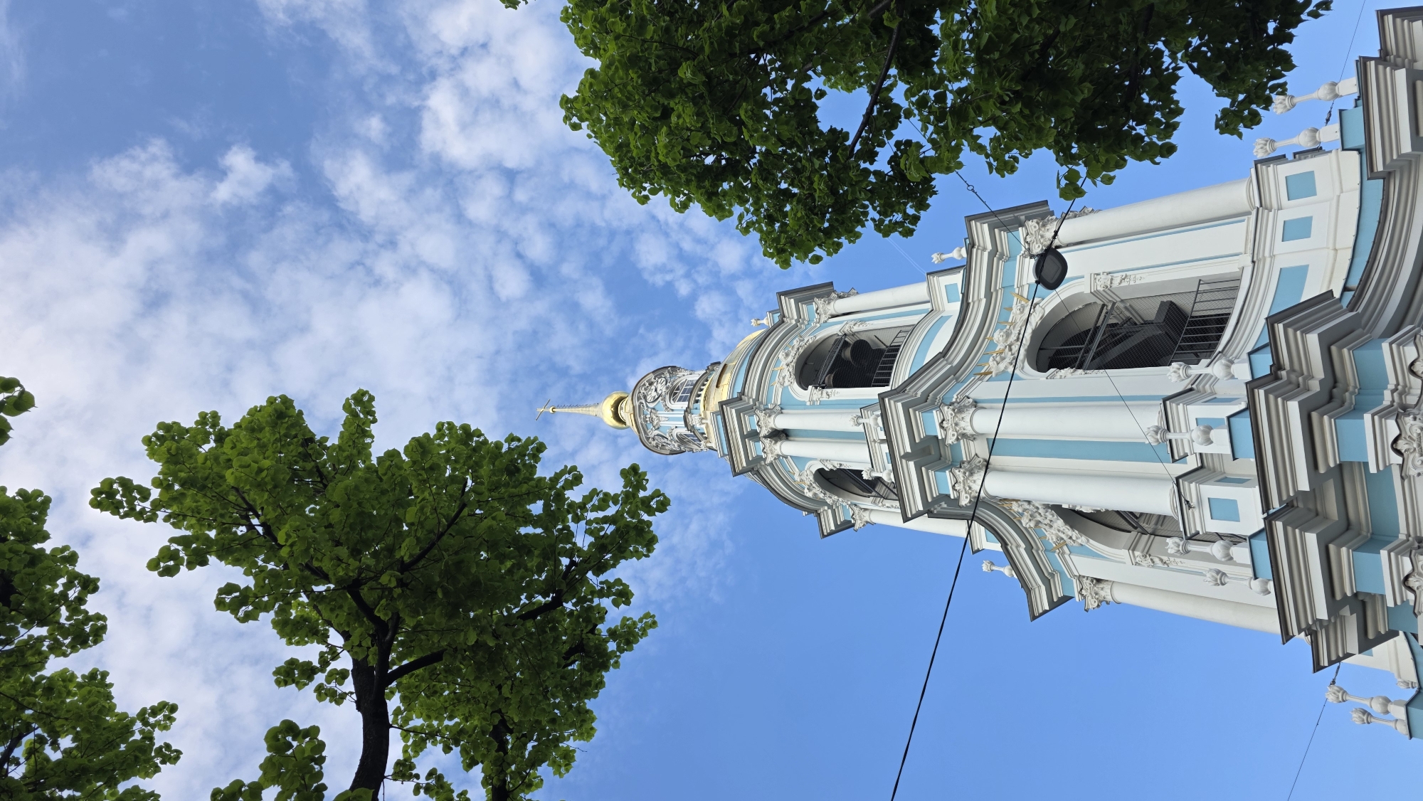 A low-angle view looks up at a blue and white baroque-style bell tower with a gilded spire in Saint Petersburg, Russia. The bell tower is framed by lush green tree branches at the lower left and upper center of the image. The sky is mostly clear blue, with scattered white clouds above and to the left of the tower. A dark, thin wire extends diagonally across the lower right of the frame, obscuring part of the tower.