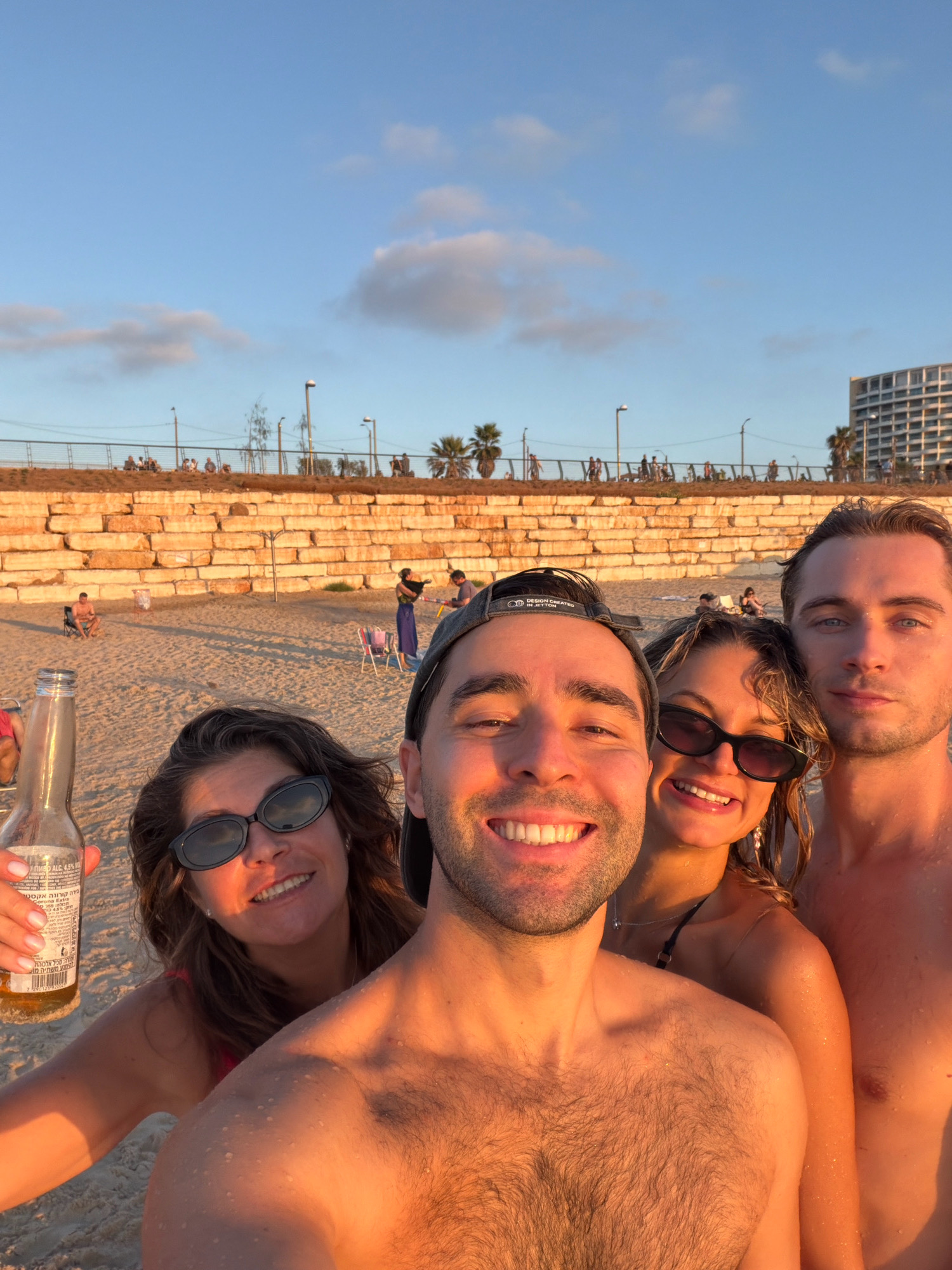 The image depicts a group of four people taking a selfie on a beach, likely in Herzliya, Israel, given the context of the provided crops showing the cityscape and building structures. The foreground is dominated by the faces of two men and two women. The man in the front center, wearing a baseball cap, is smiling widely, his chest bare and glistening with water droplets, indicating a recent swim or a humid environment. To his left is a woman, also smiling, wearing dark oval sunglasses and a pink swimsuit, holding a beer bottle in her left hand. To the right of the central man is a woman with blonde, wet hair, smiling, wearing sunglasses and a black bikini top. Further to the right is another man, with fair skin and blue eyes, shirtless, also appearing to be wet.
In the background, the sandy beach stretches out, with scattered individuals and beach chairs. A prominent feature is a large, segmented wall constructed from large, rough-hewn stones. Beyond the wall, a promenade or walkway is visible, lined with railings, streetlights, and palm trees, with a large modern building complex visible in the distance, consistent with the urban coastline of Herzliya. The sky is a clear blue with scattered, wispy clouds, and the lighting suggests it is late afternoon or early evening, with the warm golden hues of sunset casting a glow on the scene.