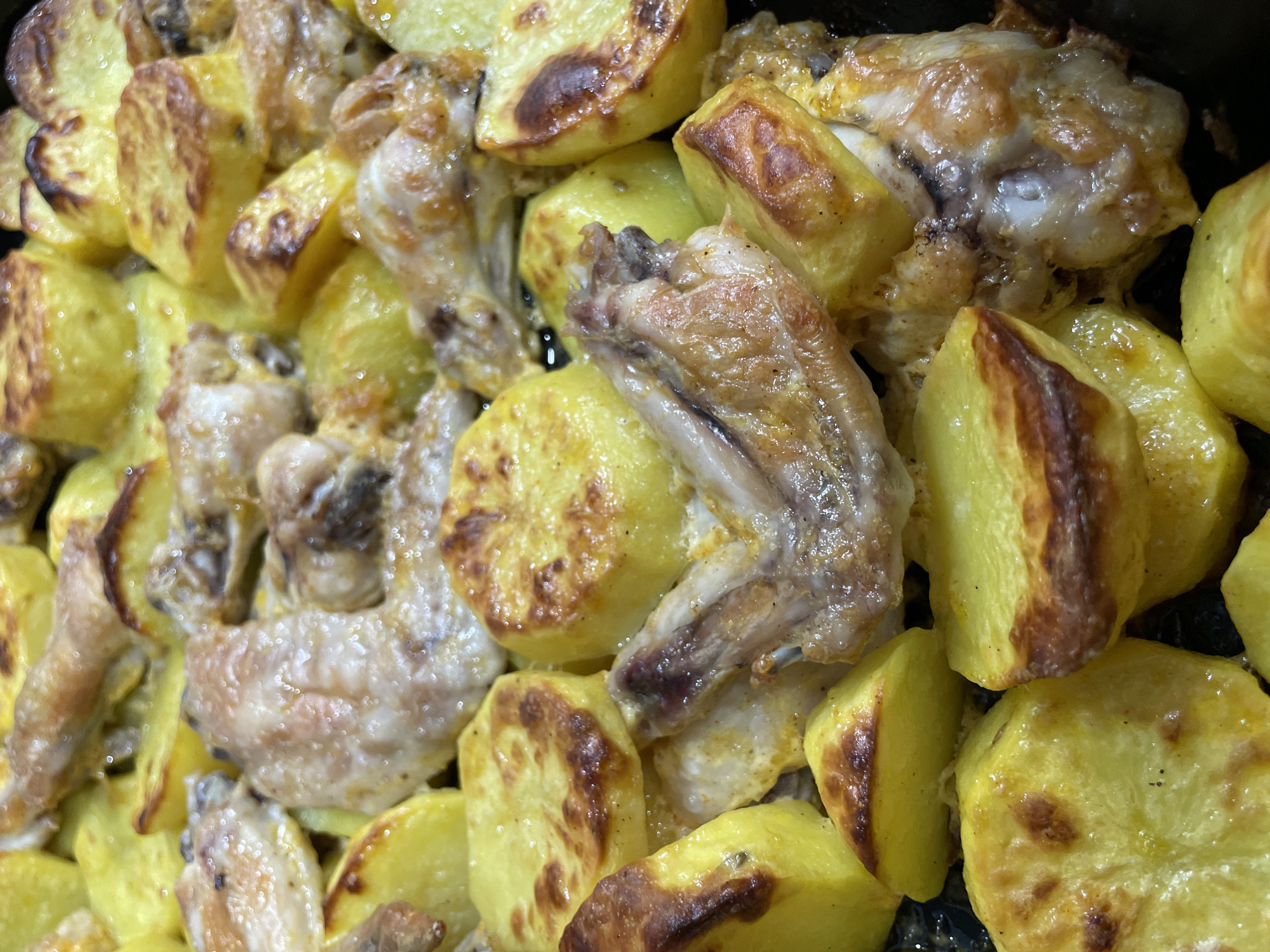 The image depicts a close-up of baked chicken wings and potato wedges. The chicken wings are light brown in color with visible skin texture. The potato wedges are yellow with browned edges and surfaces. The food appears to be arranged in a single layer, likely within a baking pan. The background is dark. No information about the location (Izhevsk, Russia) is discernible from the visual content.