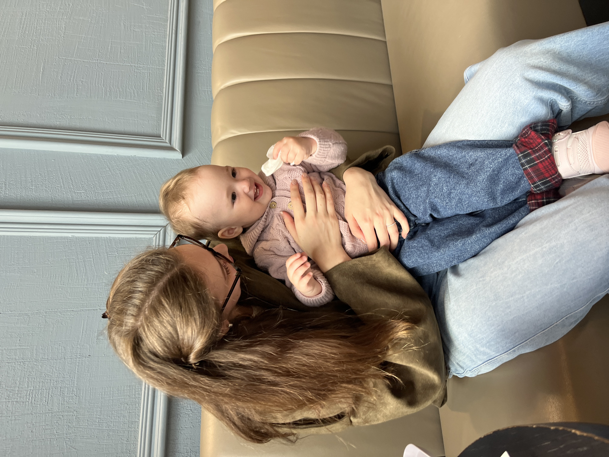 A woman with long brown hair and glasses is holding a baby on a couch. The baby, wearing a pink sweater and denim pants with red and black plaid accents, is smiling and holding a white object. The couch is light brown, and the background features a textured gray wall. The location is Shushary, Russia.