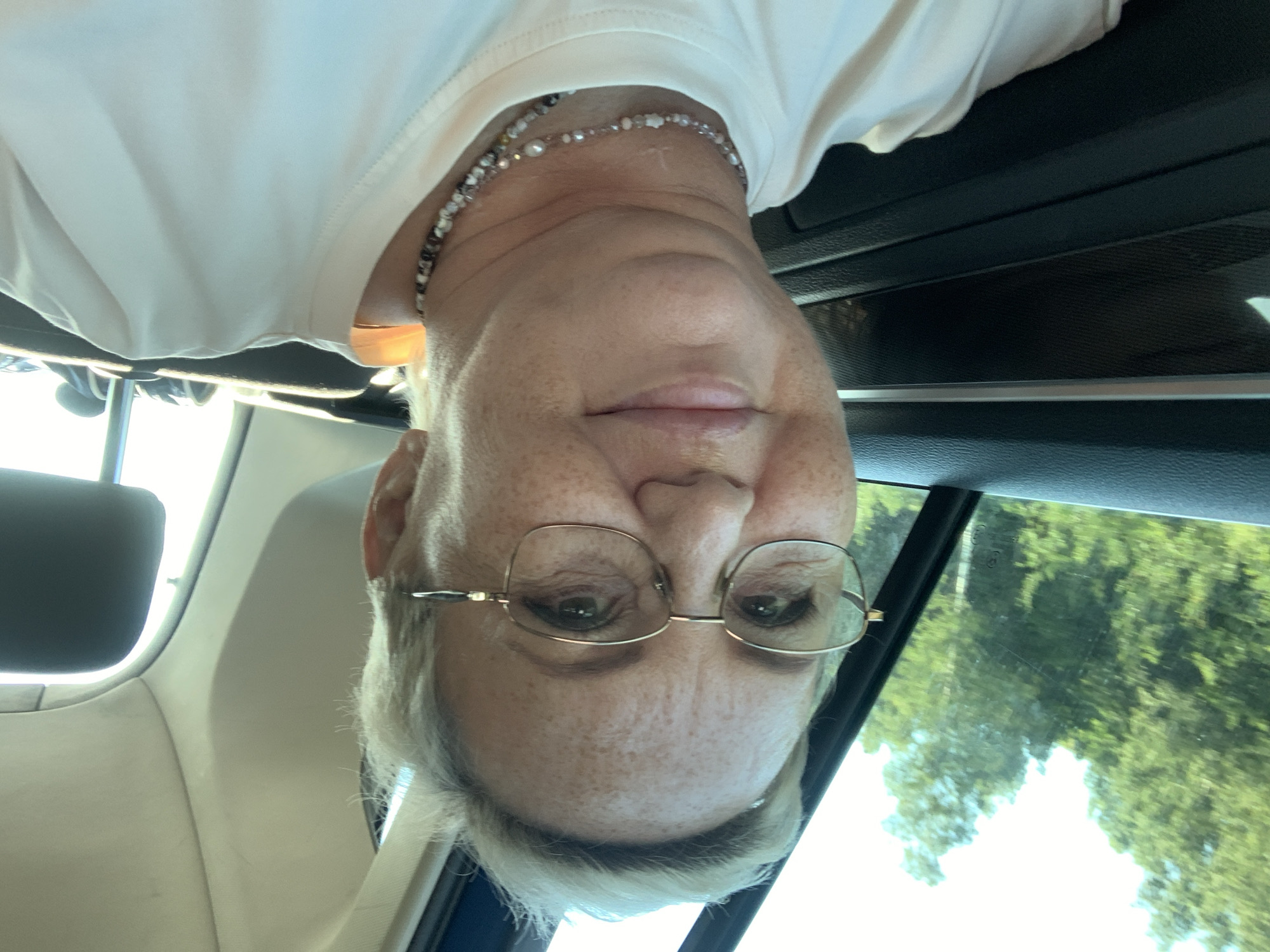 The image is a selfie of a Caucasian woman with short blonde hair and freckles, wearing glasses and a light-colored t-shirt with a beaded necklace. She is in the passenger seat of a car, with trees and a bright sky visible through the window. The interior of the car, including the dashboard and headrest, is also visible. The image does not provide any information about the city of Nizhnie Yaki, Russia.