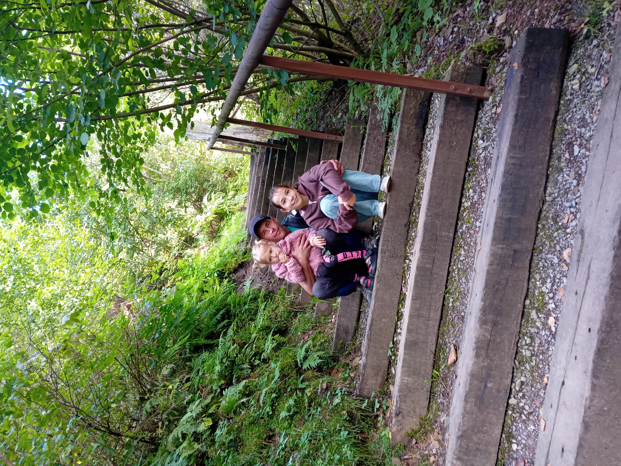 In Zuberec, Slovakia, a man and two young girls are photographed on a wooden staircase leading through a lush, green forest. The man is seated, holding the younger of the two girls on his lap. The younger girl, wearing a pink hooded jacket, has blonde hair and is looking towards the camera. Next to them, the older girl, with dark hair, is sitting with her legs crossed and her arms around herself. She is wearing a brown hooded jacket and blue jeans. The staircase is made of thick, weathered wooden beams, with a metal handrail on the right side of the frame. The forest is dense with green foliage, including ferns and trees, with sunlight filtering through the canopy. The ground surrounding the stairs is covered with gravel and fallen leaves. The overall scene depicts a family enjoying an outdoor excursion in nature.