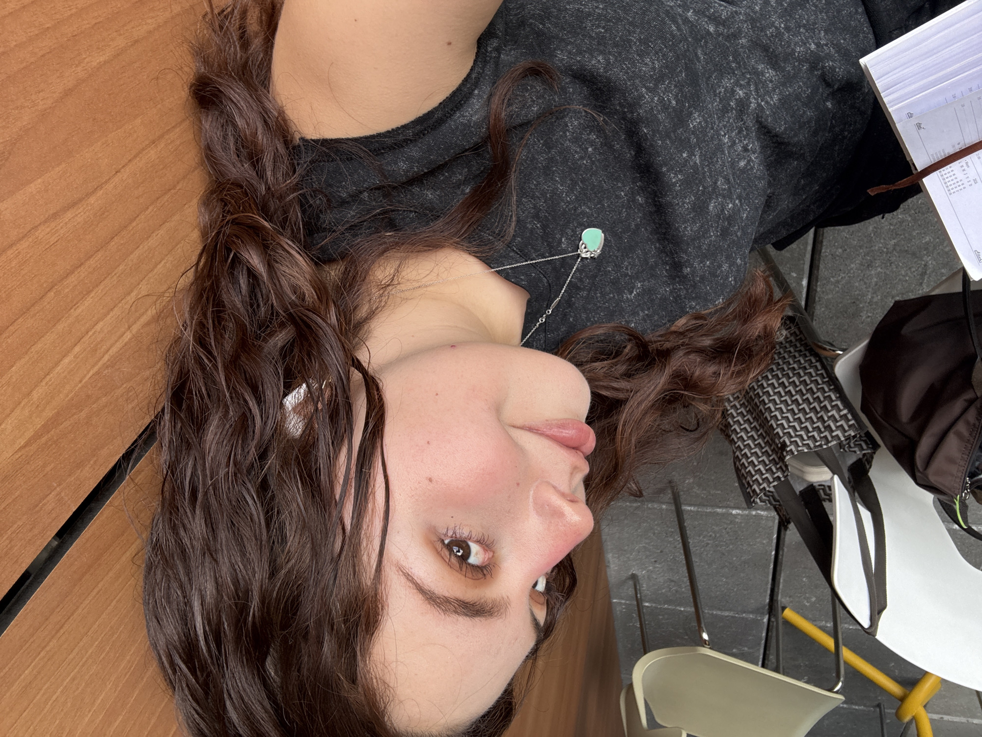 The image captures a close-up, slightly rotated view of a young adult female, likely lying or leaning on a surface. Her long, dark brown, wavy hair frames her face, with some strands appearing damp or styled. She has fair skin, brown eyes, natural eyebrows, and light-colored lips, with a subtle hint of blush on her cheeks. Her expression is calm and relaxed, with a soft, almost imperceptible smile.
She is wearing a dark gray, short-sleeved t-shirt with a slightly mottled texture. Around her neck is a delicate silver chain necklace featuring a light blue or turquoise oval-shaped pendant. A silver ear cuff or small earring is visible on her left ear.
The setting appears to be an outdoor or semi-outdoor cafe in Mexico City. To her left, her hair rests on a smooth, light brown, wood-patterned table surface. To her right, partially visible, is an open book or notebook with white pages and a brown bookmark. The text on the pages is largely illegible, but some lines and numbers, including "21:00", can be faintly discerned. Adjacent to the book is a dark brown or black bag.
In the background, two chairs are visible. One is light beige/cream with metal legs, while the other features a white seat and a dark, geometric patterned (possibly houndstooth) backrest, also with metal legs. The flooring beneath the chairs is a textured gray, possibly paved or tiled. The lighting suggests it is daytime, but there are no harsh shadows, indicating either overcast conditions or diffused lighting.