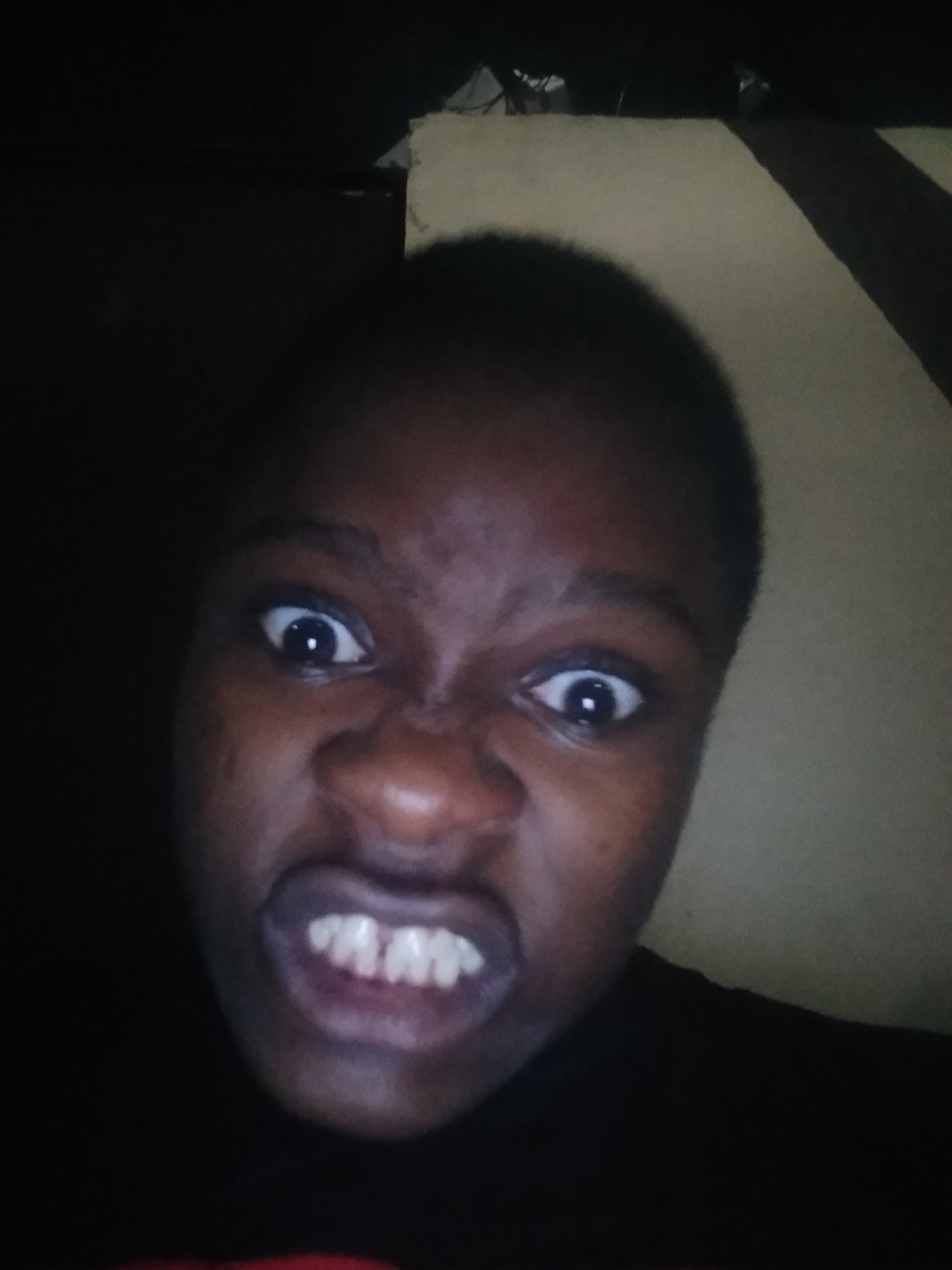 A close-up, dimly lit photograph shows a person with dark skin, making a face with wide eyes, furrowed brow, and clenched teeth. The background is partially visible and appears to be a plain wall. The location and any actions or interactions cannot be determined from the image. The location in Shangisha, Nigeria cannot be confirmed or denied, as there is no information from the image.