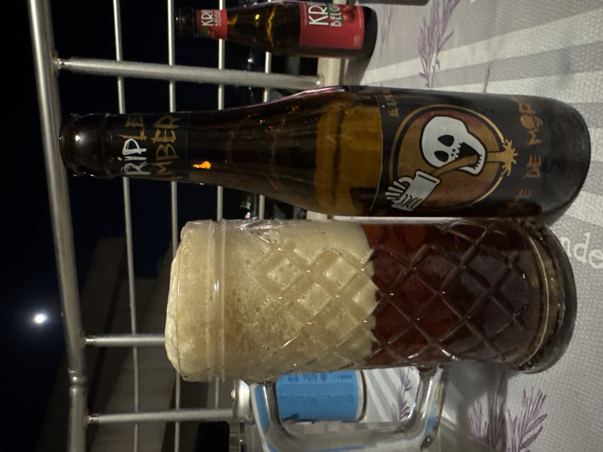 The image shows a beer glass filled with dark beer and topped with a thick head of white foam, sitting on a table or surface. Next to the glass is a dark brown beer bottle with a label featuring a white skull graphic, the label also says "NIE DE MOR". The bottle is partially obscured by metal bars. Above the bottle are other bottles with visible labels. The background includes a glimpse of a night sky and a bright moon. Based on similar images, this is likely taken at a restaurant or bar in the city of Budva, Montenegro.