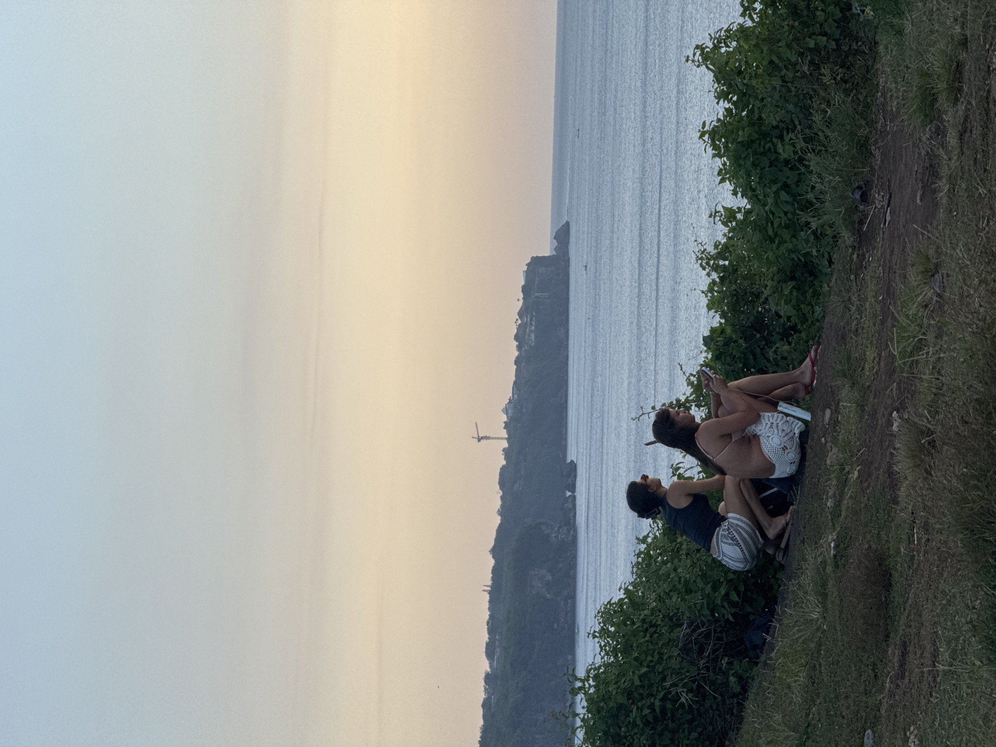 Two individuals are positioned on a grassy, uneven cliff overlooking the ocean and a rugged coastline. The sky is a gradient of light blue to soft yellow, suggesting dawn or dusk. A construction crane is visible on a distant headland. The woman is sitting with legs crossed, using a mobile phone, wearing a light-colored top and shorts. The man sits beside her, also facing the view. They are positioned amid greenery and vegetation. The location is likely in the city of Kuta Selatan, Indonesia.