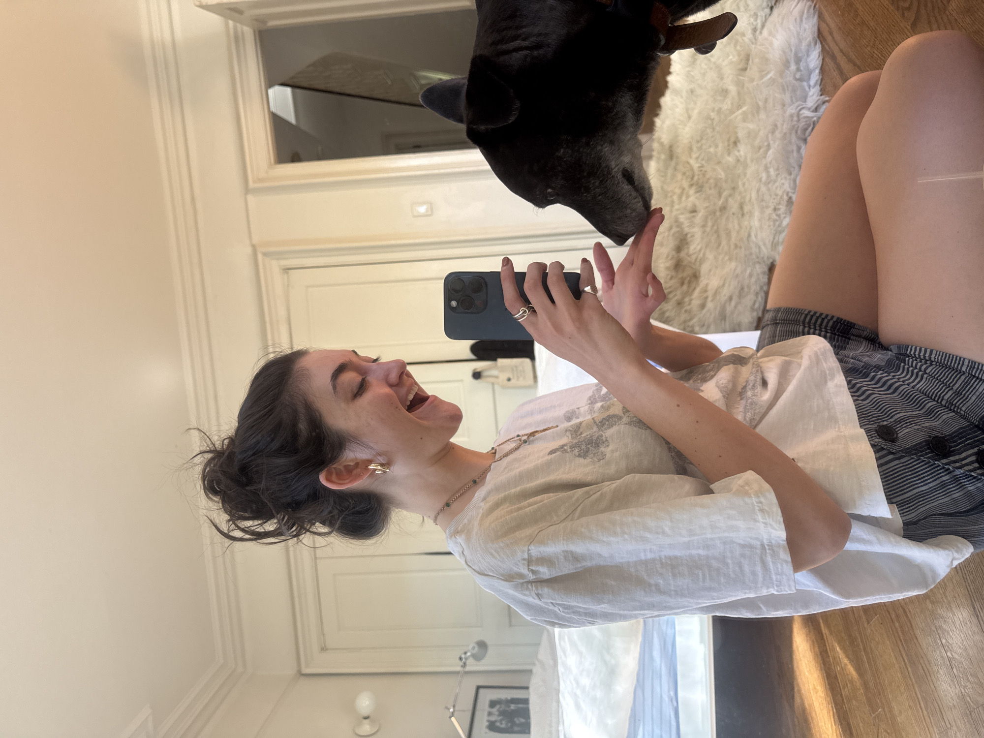 A young woman is captured mid-laugh, lying down on a wooden floor as she interacts with a black dog. Her dark hair is styled in a messy bun, and she wears a light-colored, possibly linen, t-shirt and striped shorts. She accessorizes with several rings on her fingers, earrings, and a delicate necklace. In her hands, she holds a dark smartphone, identifiable by its triple-lens camera array, with her fingers playfully extended towards the dog.
The black dog, a medium to large breed, leans down, its nose gently touching the woman's outstretched fingertips, signifying a moment of affection or play. The woman's wide-open mouth and joyful expression convey clear happiness and warmth.
The setting is a brightly lit indoor room, likely a bedroom or living space, characterized by white walls with elegant decorative molding. A plush, white, shaggy rug lies on the wooden floor beside the subjects. Reflections in a large mirror behind them hint at a bed and other furnishings. The presence of natural light suggests it is daytime. There is no visible text in the image. The scene encapsulates a heartwarming and candid interaction between a pet owner and their dog.