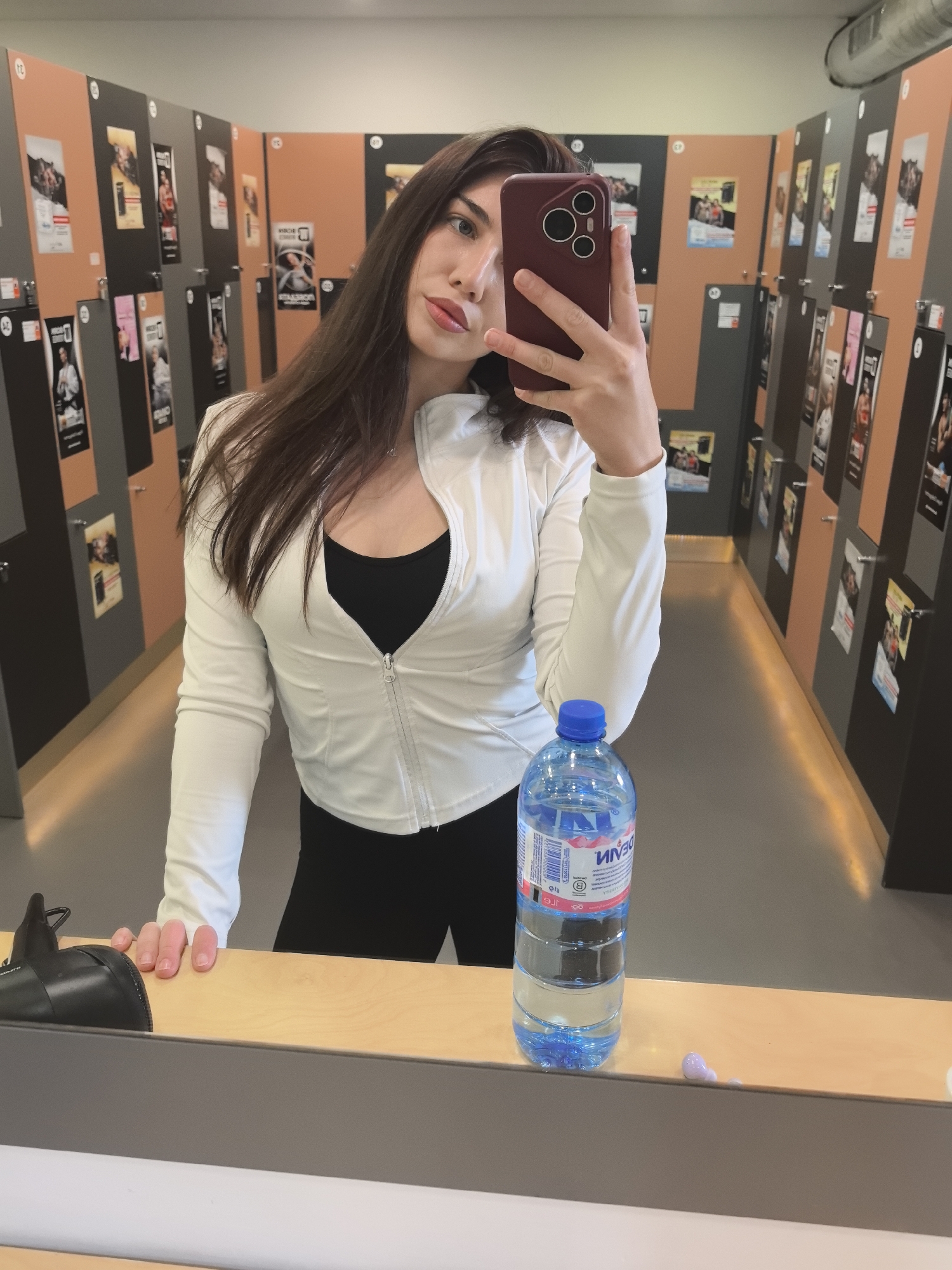 A young woman is captured taking a mirror selfie in what appears to be a locker room or changing area in Stara Zagora, Bulgaria. She stands centrally, holding a dark red smartphone with visible camera lenses in her right hand, obscuring part of her face. Her long, dark brown hair falls over her shoulders. She is wearing a white, long-sleeved, zip-up athletic jacket over a black top, paired with black leggings.
The background consists of rows of lockers or storage units, alternating between brown and dark grey panels, extending down a hallway. Numerous posters and flyers, featuring images of people and text, are affixed to these units. Some numbers, like "13", "12", and "1", are visible on the lockers. Mirrored text on one poster appears to show Cyrillic characters, possibly "МАТАЛ" or "МАТАБОГ".
In the foreground, a light wooden counter extends horizontally across the bottom of the image. On the counter, to the left, rests a black object resembling a hair dryer. To the right, a clear plastic water bottle with a blue cap is visible. The label on the bottle clearly shows "МИНАЛ" (MINAL) in Cyrillic script, mirrored, confirming the Bulgarian context. A small, pinkish-purple smudge or drop is also seen on the counter surface near the bottle.
The woman maintains a neutral, posed expression. The lighting suggests an indoor setting with overhead illumination.