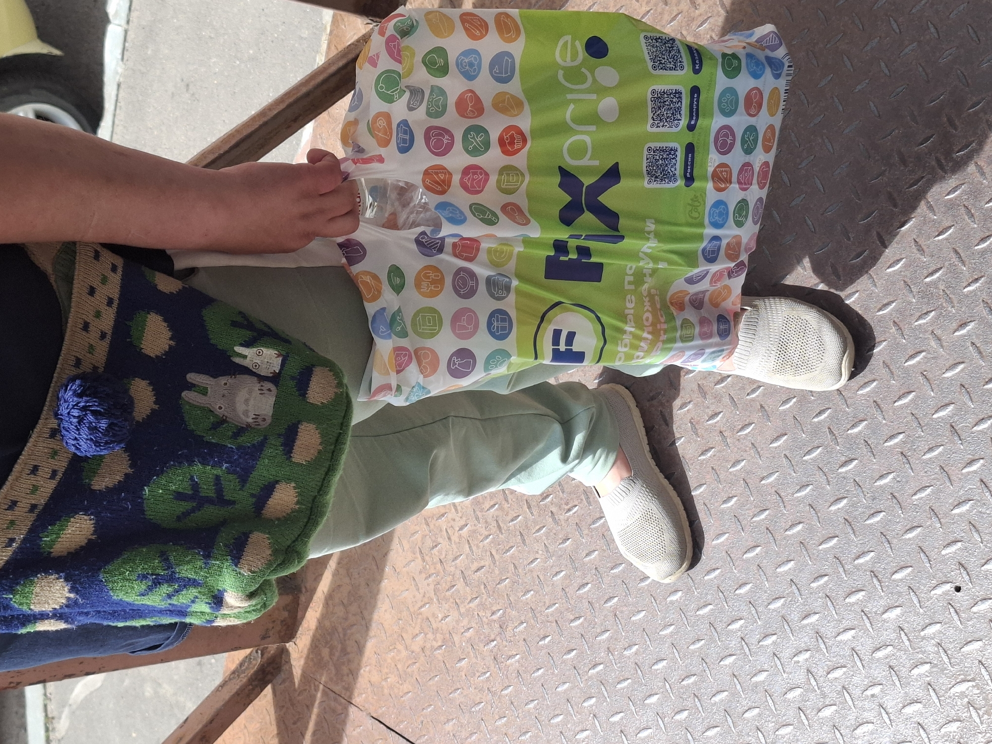 A person is standing outdoors on a metal grate floor, holding a green and white plastic shopping bag. The bag has colorful circles with icons on it and the logo "Fix price" is prominently displayed. The person is wearing light green pants and light-colored slip-on shoes. A knitted bag with a Totoro design is slung over their shoulder. The background shows a blurred view of a building and a car wheel.