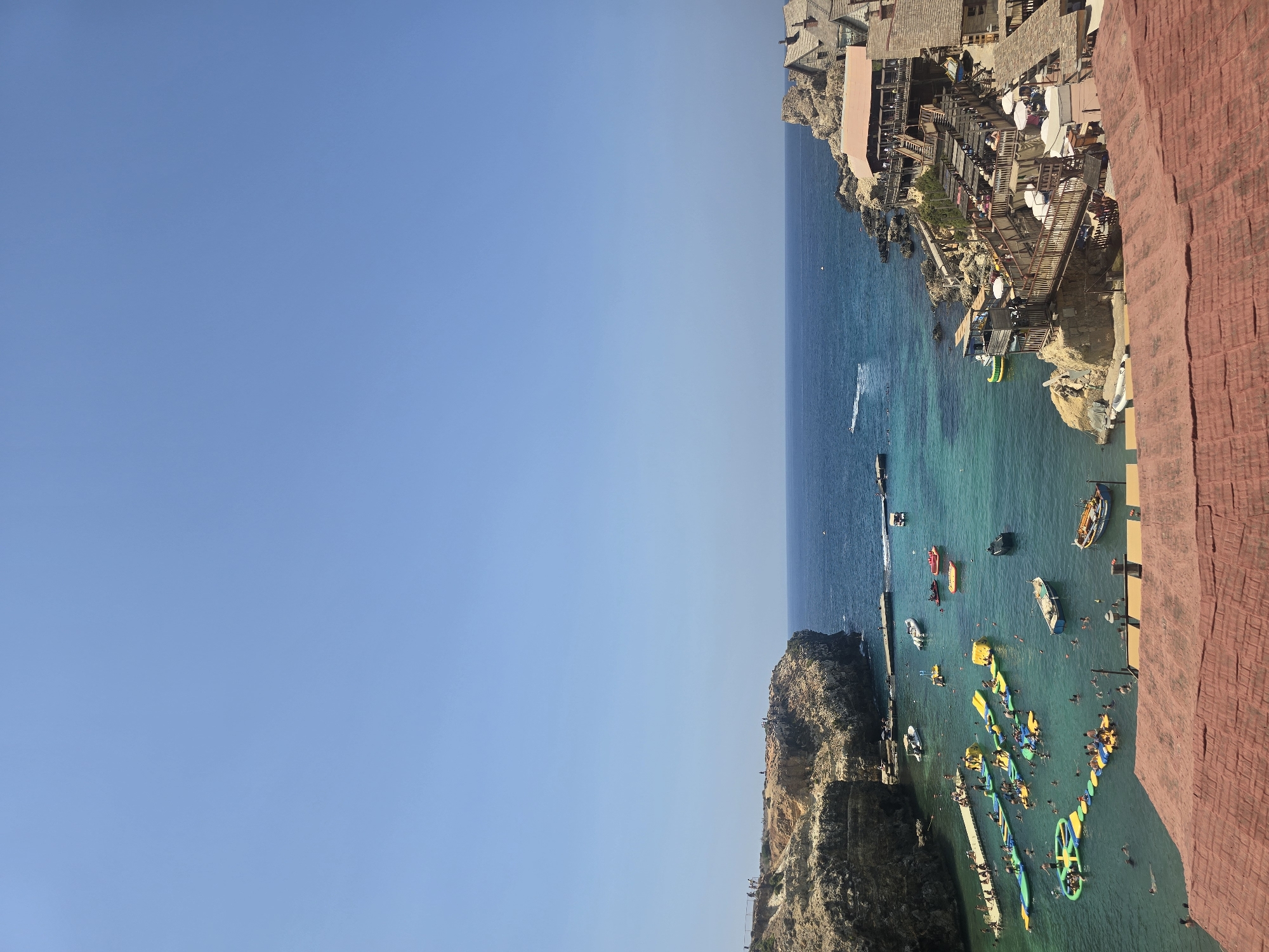 The image captures a vibrant scene at Popeye Village, also known as Anchor Bay, in Mellieħa, Malta. The crystal-clear turquoise water of the bay is filled with various activities. In the foreground, a section of a red-tiled roof is visible, suggesting the vantage point is from a building overlooking the bay.
Dominating the left side of the bay are inflatable water structures, including slides and obstacle courses, with numerous people enjoying them. Several boats are moored in the bay, some small and white, others larger and colorful. A jet ski leaves a white wake in the deeper blue water towards the open sea on the left.
The right side of the bay features the iconic Popeye Village, with its charming wooden structures built into the rocky coastline. These buildings, designed to resemble the cartoon's fictional village, house restaurants and entertainment areas, with people seated at tables and walking on wooden walkways and stairs.
Rocky cliffs form the backdrop to the scene, with a few people visible on the edge of the cliff on the left. The sky is a clear, bright blue, indicating a sunny day. The overall impression is one of a lively tourist destination on a beautiful Mediterranean day.