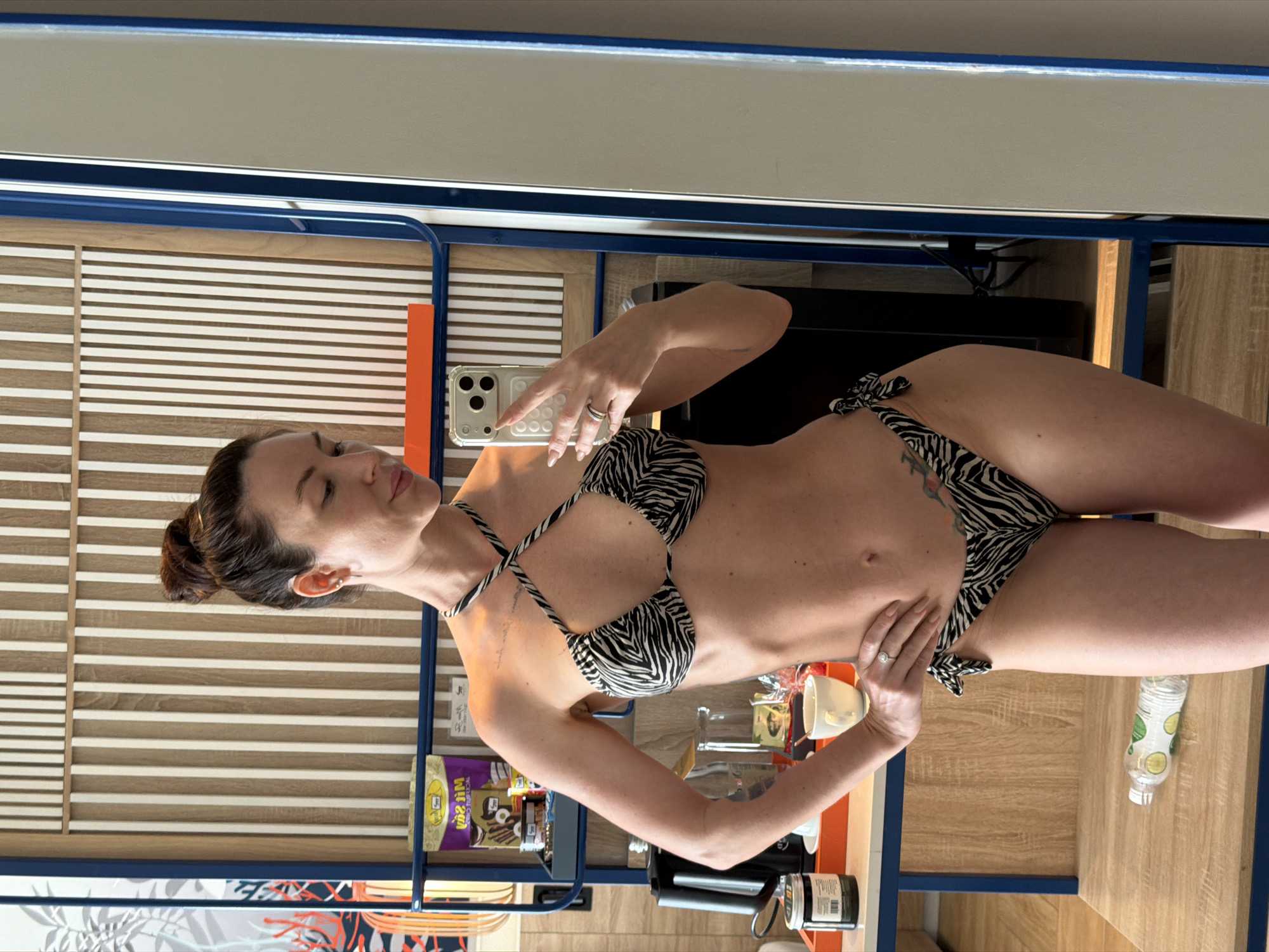 A woman with her hair in a bun is taking a mirror selfie in a room. She is wearing a black and white zebra-print bikini. She is standing with one hand on her hip. In the background, there is a shelf with various items, including snacks, drinks, and a kettle. The room appears to be a hotel room. The lighting is bright. The location is not specified in the image or any accompanying information.