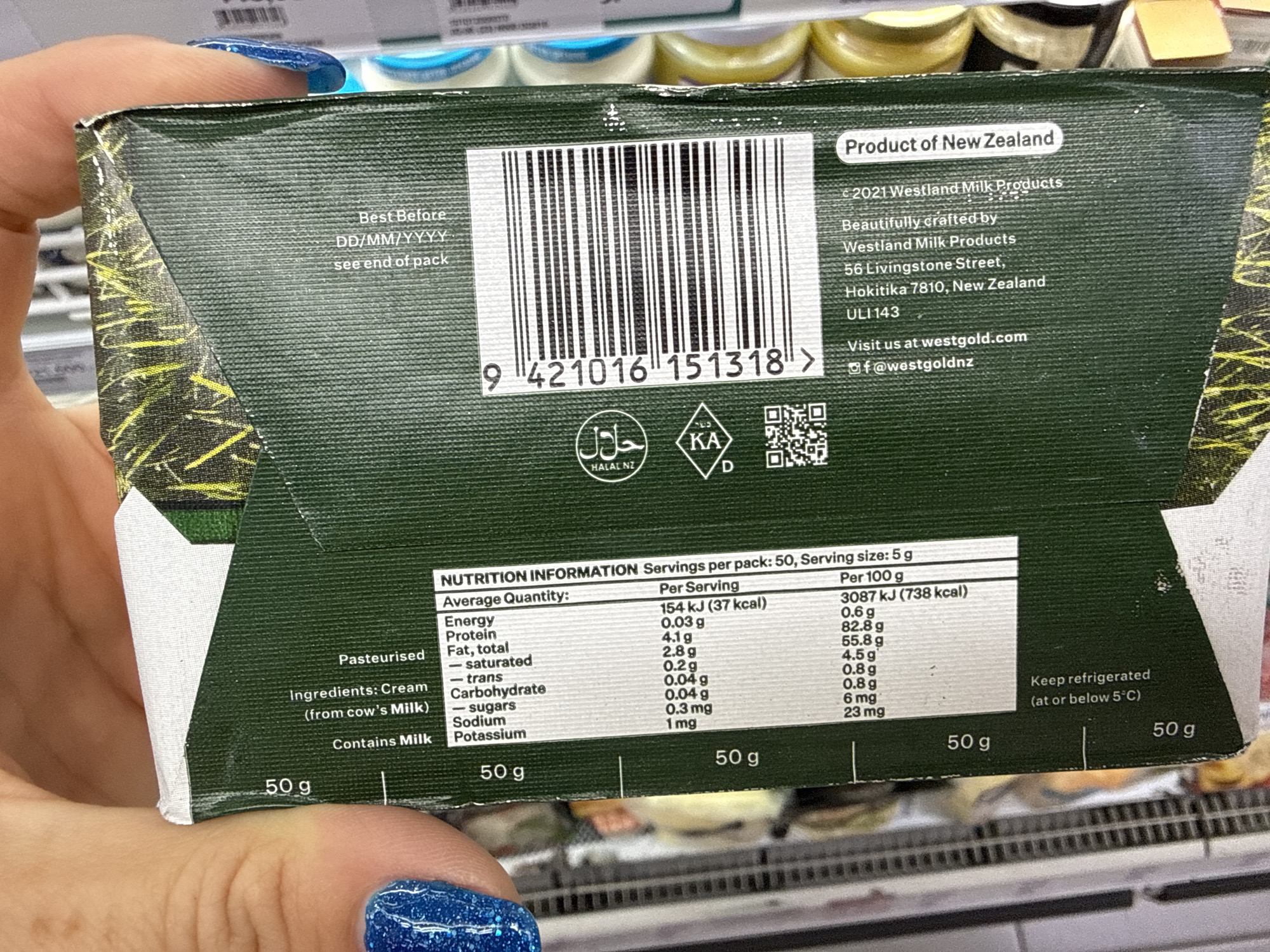 The image shows the side panel of a green cardboard box, likely packaging for a food product. The panel features nutritional information in English, including serving size, energy content, and macronutrient breakdown per serving and per 100g. It also lists ingredients, stating "Cream (from cow's Milk)" and "Pasteurised." A barcode is prominently displayed, along with logos for "HALAL NZ" and "KA D." There's also a QR code and website address, "visit us at westgold.com," and an Instagram handle "@westgoldnz." The text "Product of New Zealand" is visible in an oval shape. The box appears to have been handled, as indicated by the presence of a finger with blue glitter nail polish at the top left edge and some creasing on the cardboard. The background is blurred, but suggests a retail environment with shelves of products. The location is not specified in the image.