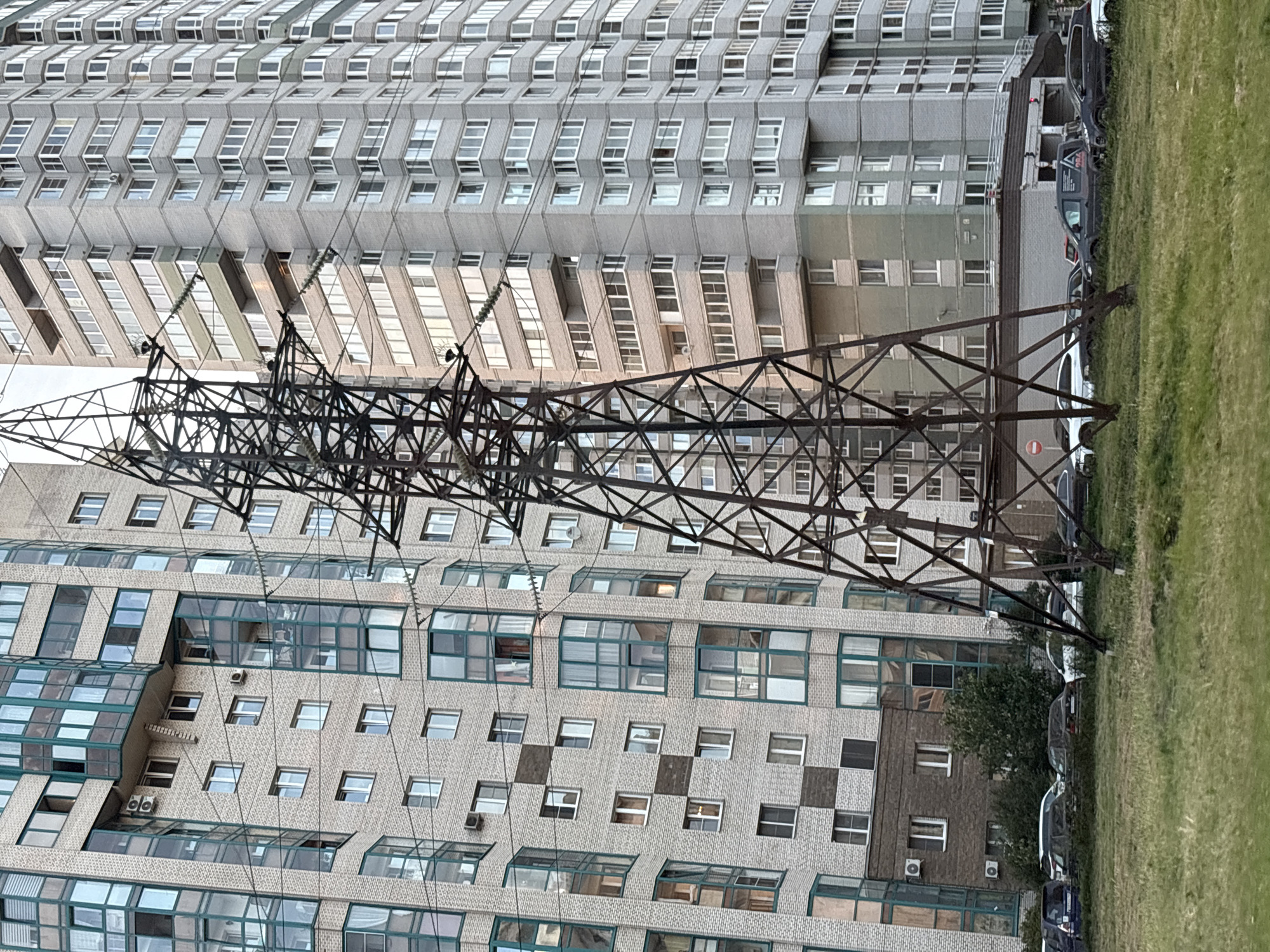 The image depicts a view of two tall residential buildings and a power line tower in Saint Petersburg, Russia. The buildings are constructed with light-colored brick and have many windows, some of which are framed in green. One building has a varied facade with alternating brick colors and inset balconies, while the other is a more uniform grey. The power line tower, made of dark metal, stands prominently in the foreground, with wires stretching across the frame. In front of the buildings, a patch of grass is visible, with several parked cars of various colors and models. The sky is overcast, suggesting a cloudy day.