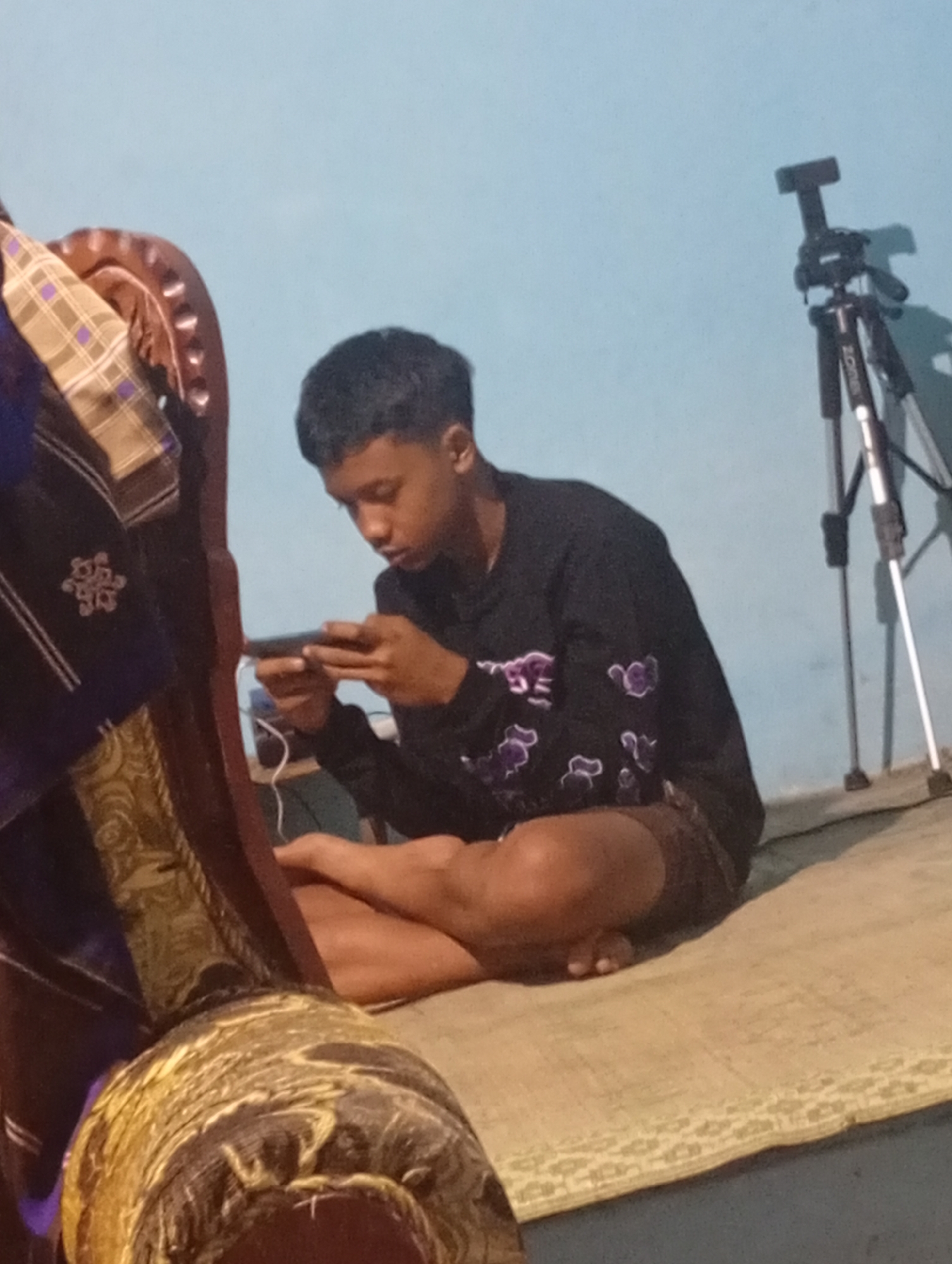 A young man is seated on a woven mat in front of a light blue wall, holding a mobile phone. He is wearing a dark long-sleeved shirt with purple floral patterns and brown shorts. His legs are crossed, and he is looking down at the phone, his thumbs actively engaged with the screen. To his left, a portion of a decorative chair with plaid and embroidered fabric is visible. In the background, a black tripod with a camera mount stands against the wall. The scene appears to be indoors, possibly in a residential setting in Mriyan, Indonesia. The lighting is dim, casting shadows on the wall.