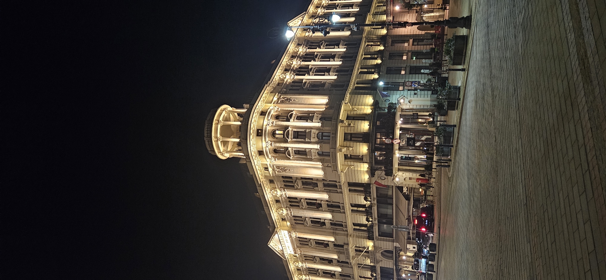 The image depicts the historic Hotel Bristol in Warsaw, Poland, at night. The ornate building, illuminated by warm yellow lights, stands prominently against a dark sky. The architecture features classical elements such as columns, arched windows, and decorative friezes. A circular dome crowns one section of the building. In front of the hotel, a paved street is visible with several cars parked and moving. Streetlights cast pools of light on the sidewalk and cobblestone-like pavement. Planters with greenery line the street, and a few people can be seen walking or standing near the hotel entrance. The scene conveys a sense of elegance and the vibrant atmosphere of the city at night.