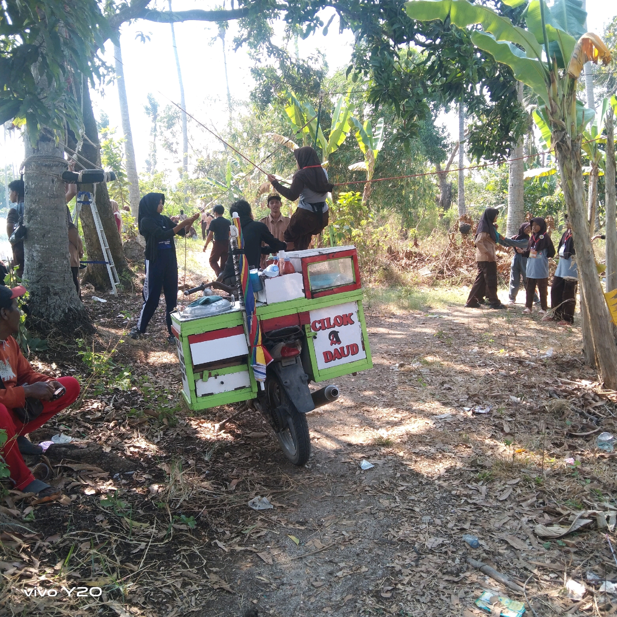 In a sun-dappled, outdoor environment that appears to be a rural or semi-rural area in Karawang, Indonesia, a group of individuals, predominantly young women wearing hijabs and uniforms, are engaged in what looks like an outdoor activity, possibly a school event or scout meeting. In the foreground, a man in a red shirt and black pants is seated on the ground, looking at his phone. Behind him, a motorcycle is parked, fitted with a green and white food cart. The cart displays the text "CILOK A DAUD!" in red lettering. Several trees with lush green foliage dominate the background, with some banana trees visible. Ropes are strung between trees, suggesting an obstacle course or recreational setup. Some individuals are holding long poles, possibly for the activity. The ground is covered with dry leaves and scattered debris, indicating a natural, unpaved setting. The overall impression is of a lively outdoor gathering amidst a tropical landscape.
