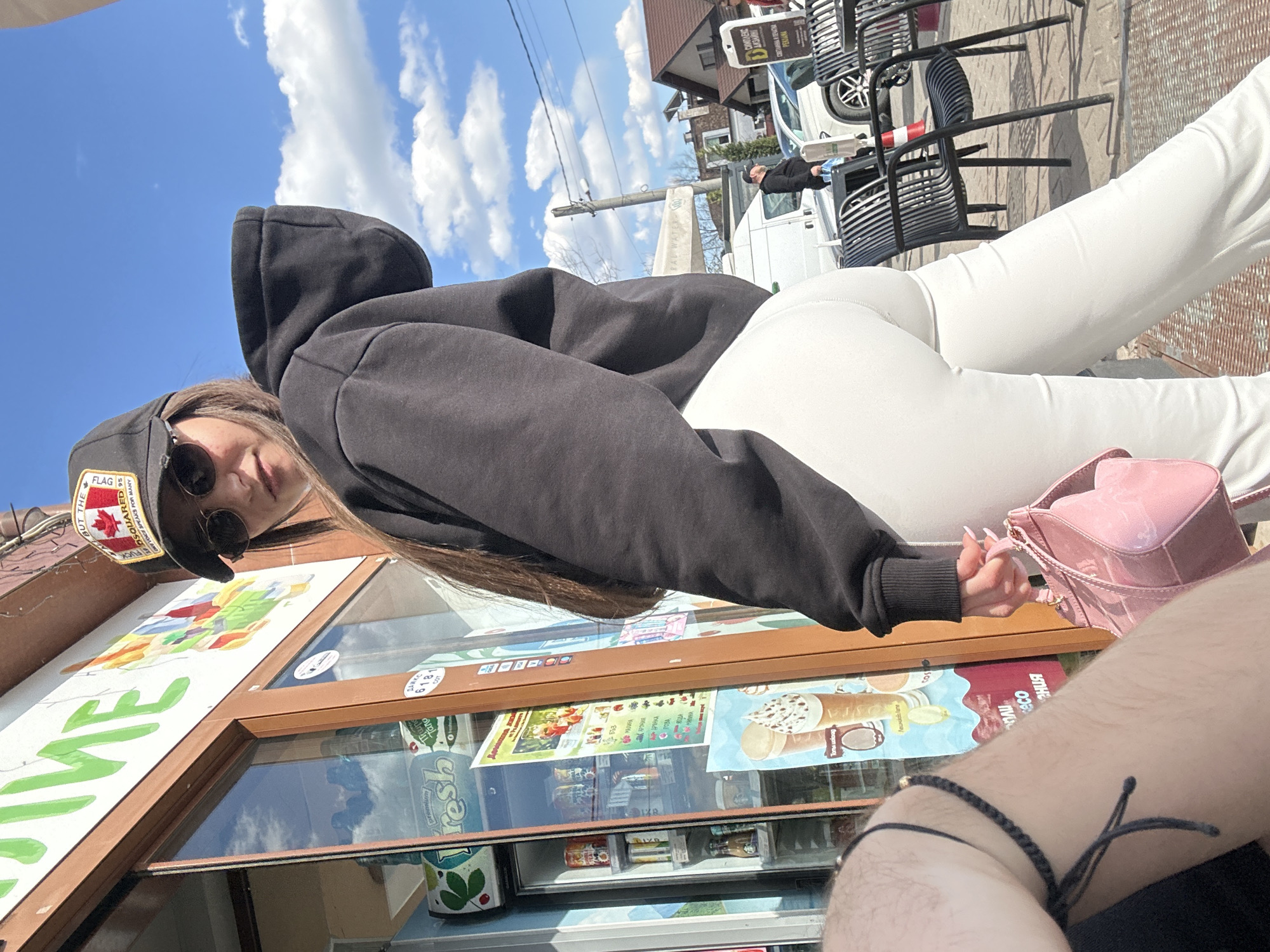A medium shot captures a person in a dark hoodie and white pants, sitting at an outdoor café in Plovdiv, Bulgaria. The individual, wearing sunglasses and a black baseball cap with a Canadian flag, is positioned in front of an ice cream shop window, which displays various advertisements and product information. A pink purse rests beside them. Seated opposite is another person, partially visible, with a dark bracelet on their arm. In the background, a street scene includes a white van, other individuals, and a row of empty chairs. The sky overhead is blue with scattered clouds. The scene is generally well-lit, indicating daytime.