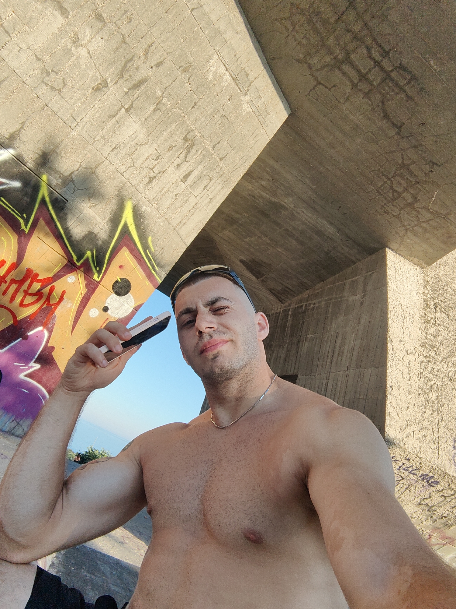 A man is taking a selfie outdoors. He is shirtless and wearing sunglasses on his head. He is holding a light-colored smartphone in his right hand up to his face. The man is smiling and appears to be relaxed. Behind him, there is a large concrete structure with graffiti on it. The graffiti is colorful, with red, yellow, and purple spray paint visible. The sky is clear and blue, and the sun is shining, suggesting it is daytime and likely warm weather. The concrete structure appears to be part of an outdoor public space, possibly an abandoned building or an urban art installation. The background shows a glimpse of water and trees, hinting at a coastal or waterside location. The man's skin has some tan lines. There is text in the graffiti, but it is not clearly legible. The overall impression is a candid and casual moment captured on a sunny day in an urban or semi-urban environment.