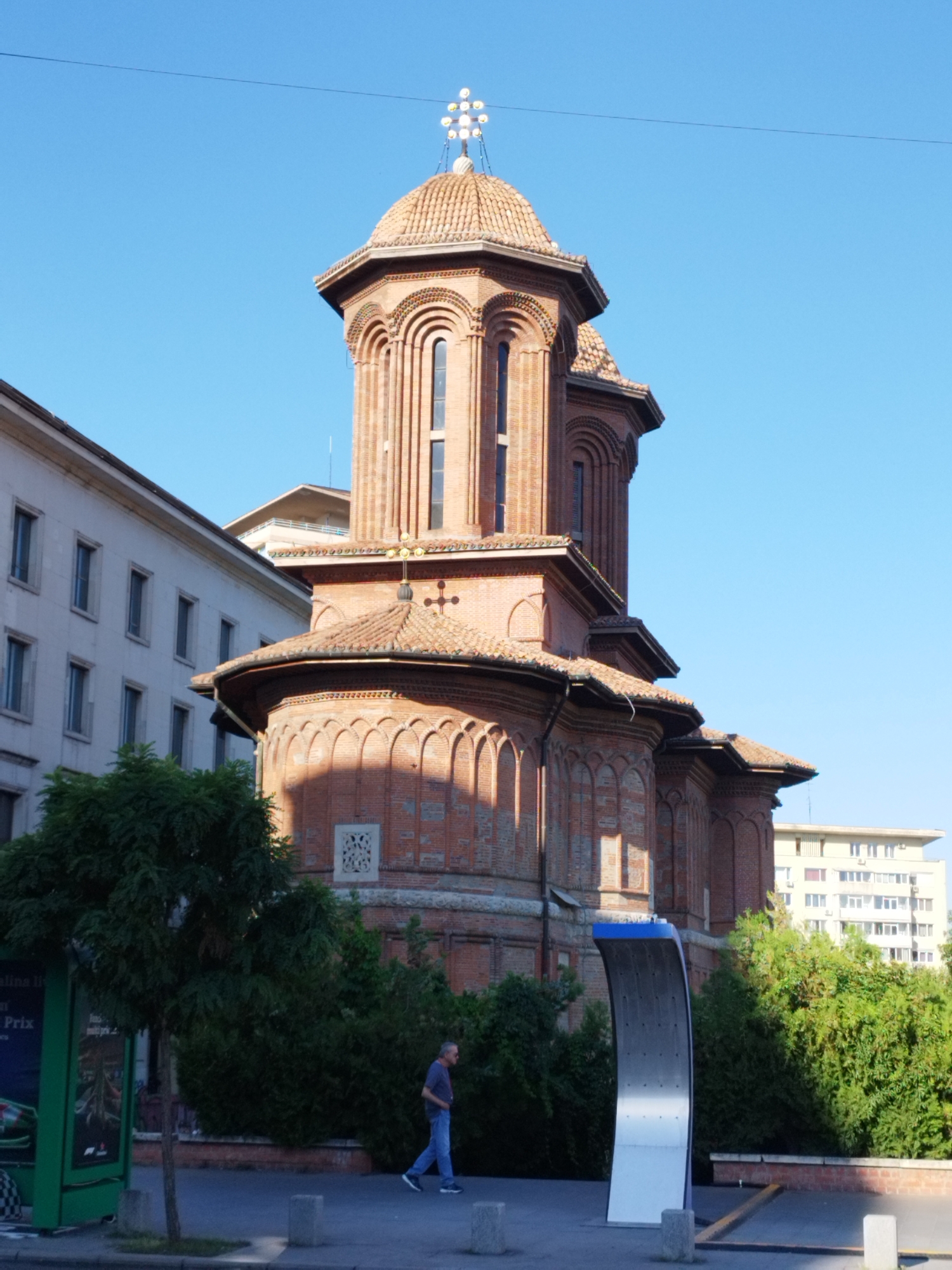 The image shows the Stavropoleos Monastery Church in Bucharest, Romania. The church is a prominent landmark made of red brick with arched windows and tiled roofs. It is a multi-tiered structure with a central dome topped with a cross. In front of the church, there is a person walking on a sidewalk. Behind the church, there are other buildings and trees, suggesting an urban environment. The sky is clear and blue, indicating it is daytime.