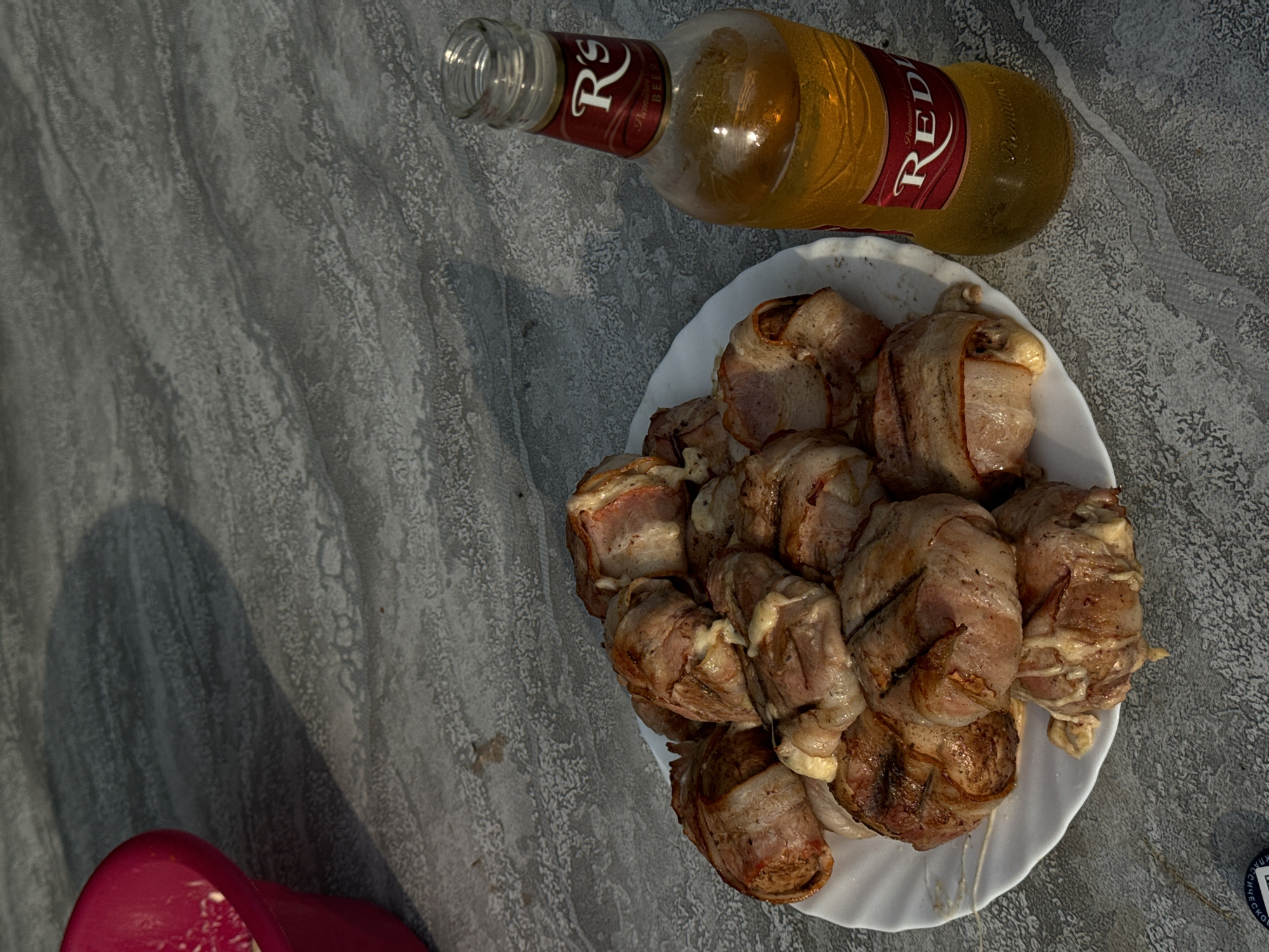 The image shows a plate of food and a bottle of beer on a textured grey surface. The food consists of several pieces wrapped in bacon, likely grilled, and arranged on a white plate. The beer bottle is a clear glass bottle with a red and gold label that reads "RED'S Premium Beer". The background is a grey, concrete-like surface with visible texture and some shadow. The scene appears to be outdoors. There is a red bowl partially visible in the upper left corner of the image, containing some white substance. The image does not contain any discernible elements that would allow for the identification of the city as Поназырево, Russia.