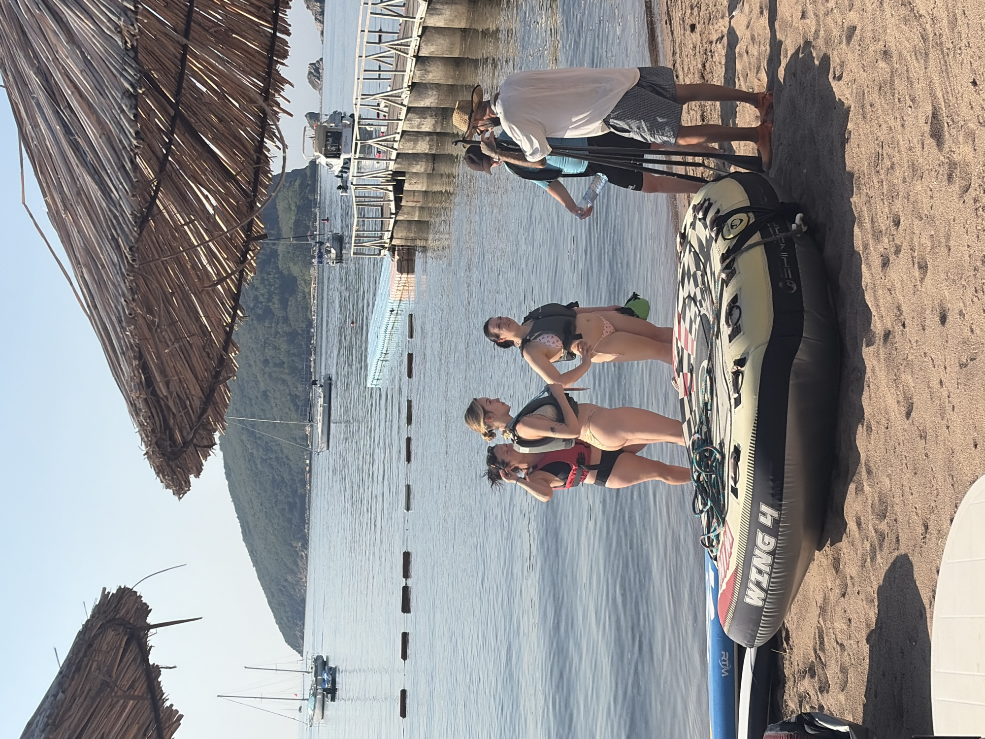 The image depicts a beach scene in Budva, Montenegro. In the foreground, an inflatable tube with "WING 4" branding rests on the sand. To the left of the tube, a blue paddleboard is partially visible. In the water, four individuals are standing at the edge of the sea. Two women are wearing swimsuits and life vests, while another woman is wearing a swimsuit and appears to be holding a green bag. A man in a straw hat and a white shirt is interacting with a man in a blue shirt and a baseball cap, who is holding a water bottle. In the background, a wooden pier extends into the calm sea, with a few boats moored nearby. Beyond the water, a lush green island rises, and sailboats are visible on the horizon. The sky is clear and blue. Two large, thatched umbrellas provide shade in the upper portion of the image.