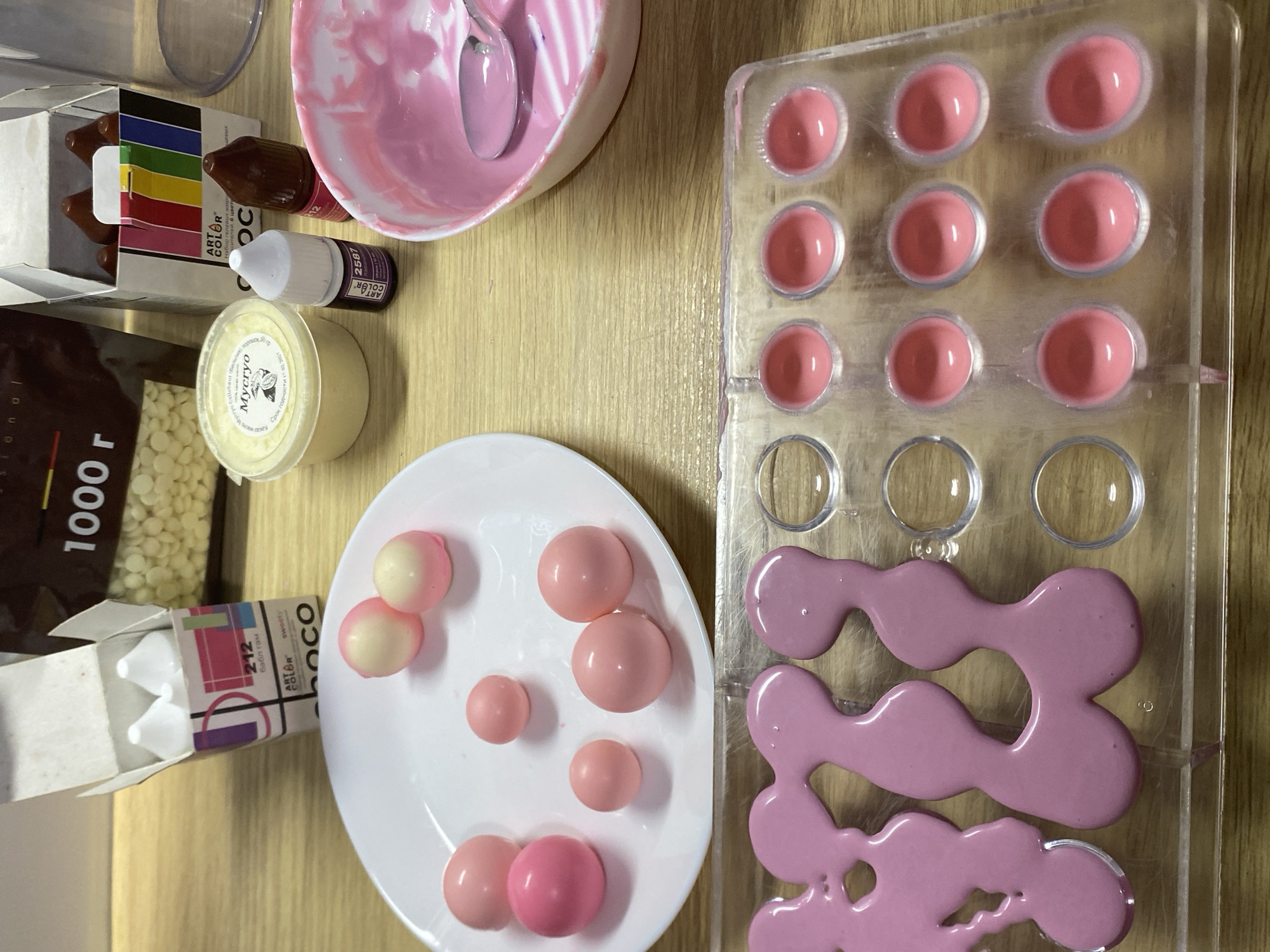 The image shows a close-up overhead view of a wooden table with various items related to candy making. In the foreground, a clear plastic mold contains pink liquid, likely chocolate or candy melt, being formed into various shapes. To the left, a white plate holds several round, pink and white candies, some solid pink and some with a lighter hue. Behind the plate, a large bag labeled "1000 г" (1000g) with white chocolate chips is visible. To the right of the bag, a box of colorful food coloring bottles, labeled "ART COLOR," stands open. Next to it, a small plastic tub with a yellow substance and a label in Cyrillic script is present, along with a small dropper bottle of purple coloring and a dark brown bottle of coloring. Further to the right, a white bowl contains pink liquid and a spoon, suggesting the preparation or application of coloring. The lighting is bright, casting soft shadows, and the overall scene suggests a home kitchen or a small confectionery workspace. There are no people or animals visible in the image, and the location within Kushchovskaya, Russia is not discernable from the visual information provided.