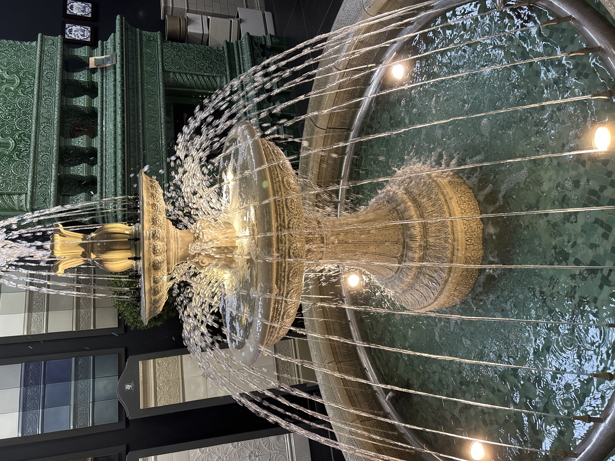 An elaborately carved, multi-tiered fountain stands prominently, actively spraying water from its various levels. The fountain appears to be made of light-colored stone or ceramic, adorned with intricate, classical-style patterns. Jets of water arc outwards from the upper and lower tiers, creating a shimmering veil, and fall into a large, circular basin below. The basin is filled with dark green water, possibly a mosaic of dark green and blue tiles, with several illuminated lights visible within the water, reflecting off the disturbed surface.
Behind the fountain, a striking architectural feature dominates the background: a richly textured wall or display composed of large, emerald-green ceramic tiles. These tiles feature embossed floral and geometric patterns, resembling ornate decorative paneling. To the left and right of this green display, and in the foreground in some sections, are multiple flat display panels showcasing various decorative tiles in different colors and patterns, including blue, white, grey, and beige, some with textured or relief designs. The setting appears to be indoors or at night, indicated by the artificial lighting that illuminates the fountain and the surrounding displays, creating a sophisticated and elegant atmosphere, likely within a showroom or design store. No discernible text is clearly visible on any of the surfaces.