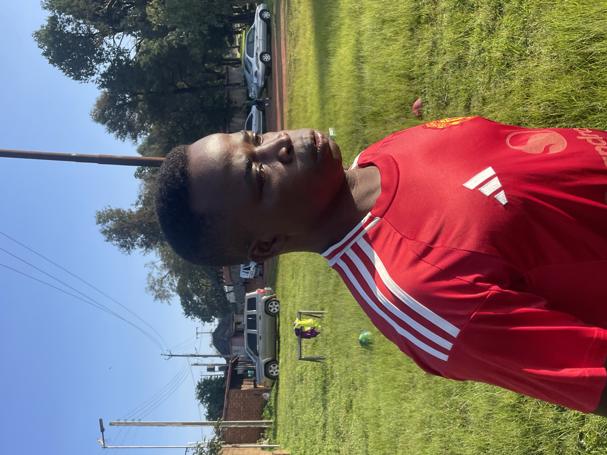 A medium shot captures a young Black male in profile, facing right, outdoors in a grassy area. He is wearing a red soccer jersey with white stripes on the sleeves and Adidas and Manchester United logos on the front. His facial expression is neutral, with his mouth slightly open. The background shows a light-colored building with a red-tiled roof, a silver SUV, and several trees. The ground is covered in green grass. The sky is a clear, bright blue. The scene appears to be set in a residential area on a sunny day.