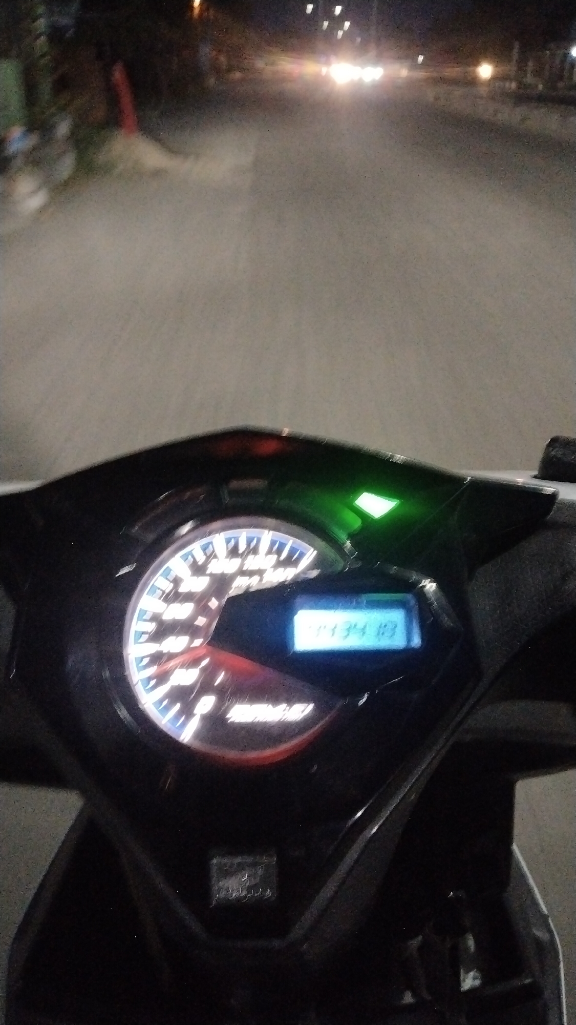 A first-person perspective of a motorcycle dashboard is captured at night on a paved road in Lhokseumawe, Indonesia. The dashboard features a speedometer with illuminated markings and a digital display showing "1134 10". A green indicator light is also illuminated. In the background, distant vehicle headlights and streetlights are visible, suggesting a moderately lit urban or suburban environment. The road surface appears wet, reflecting some of the ambient light. The surrounding area is dark and somewhat blurred, hinting at motion.