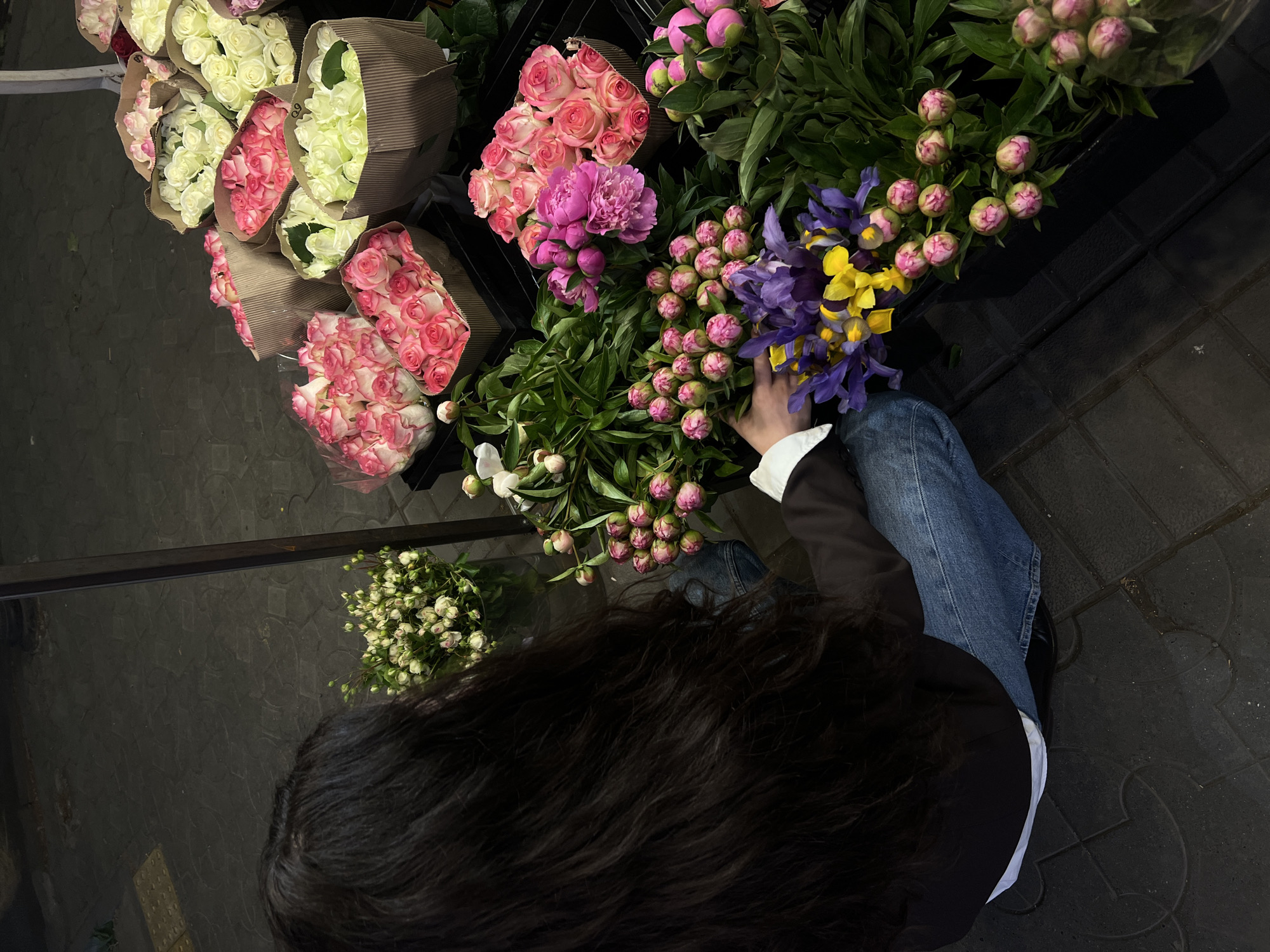 A high-angle shot depicts a woman selecting flowers from an outdoor floral display in what is likely Yerevan, Armenia. The floral selection includes bunches of white and pink roses in brown paper, as well as assortments of peonies and irises, some with closed buds. The woman, wearing a black blazer over a white shirt and blue jeans, has dark hair. The scene is set on a dark paved area.
