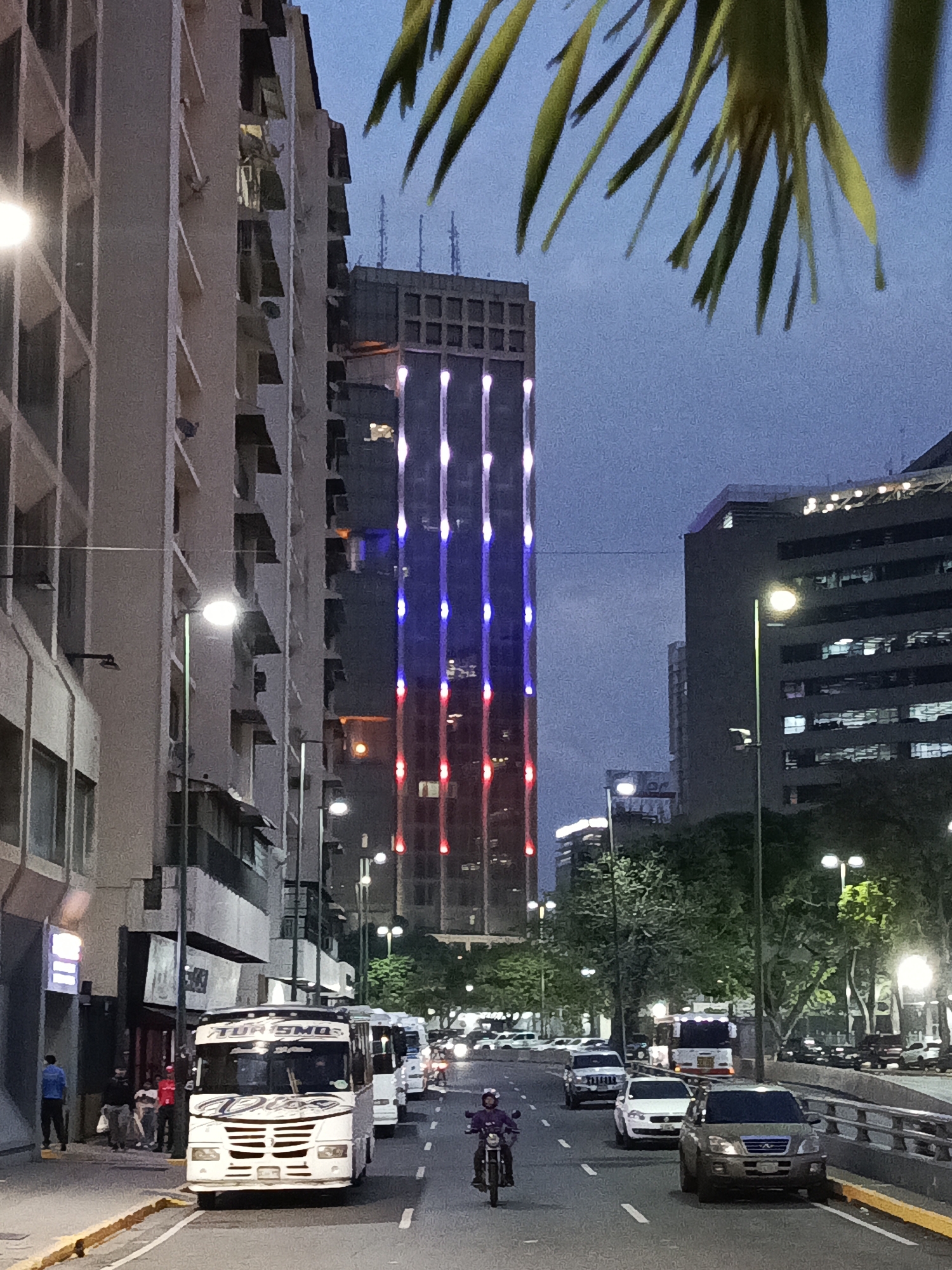 The image shows a street in an urban environment, likely at dusk or early evening. The prominent building in the background is a skyscraper illuminated with vertical bands of blue, white, and red lights, resembling a national flag. To the left is another, older building with a repetitive architectural style of projecting balconies or architectural elements. Streetlights are visible along the street. The road is active with vehicular traffic, including a motorcycle in the foreground, several buses, and other cars. There are pedestrians visible on the left sidewalk. Palm tree foliage is visible at the top right.
