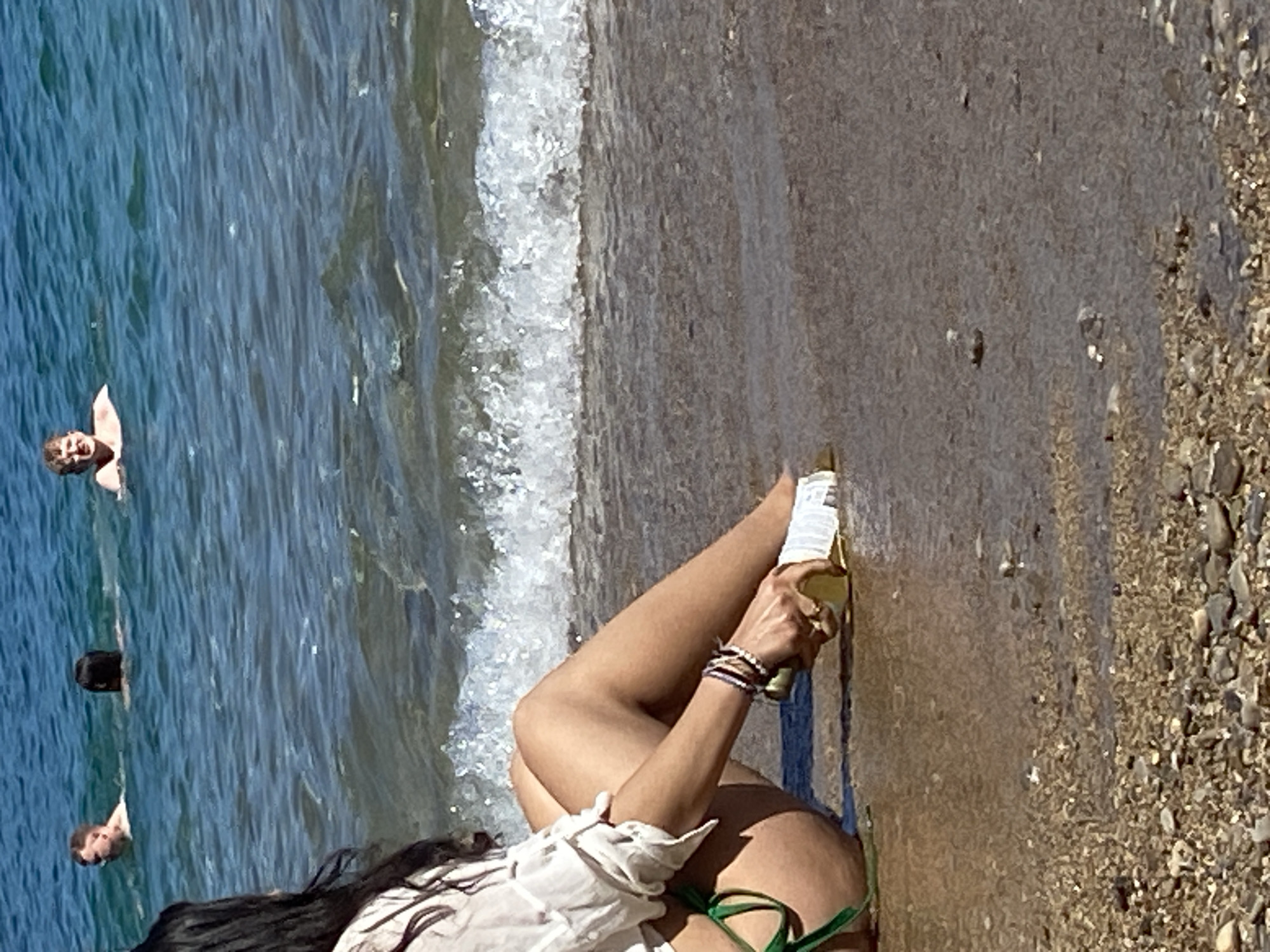 A woman is lying on a beach in Budva, Montenegro. She is wearing a white shirt and green bikini bottoms. She has long, dark hair and is wearing several bracelets on her wrist. She is holding a beer bottle. The beach is sandy with some small rocks. The sea is blue and there are people swimming in the distance.