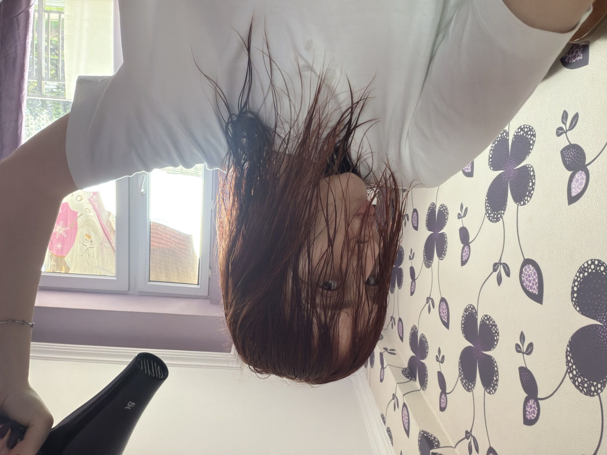 A woman with auburn hair is upside down, drying her hair with a black hair dryer. She is wearing a white t-shirt. The background shows a window with a white frame, and a white wall with a pattern of purple flowers. The location is not possible to determine.