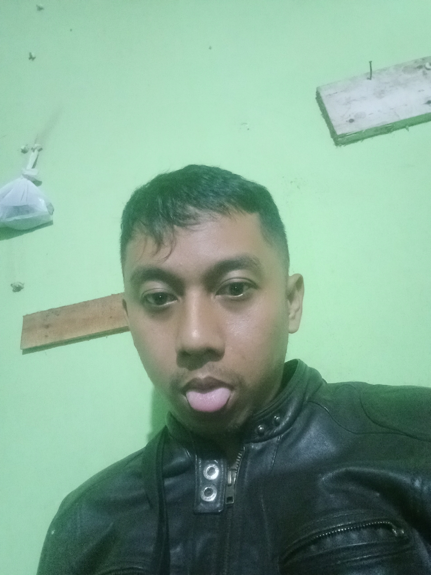 The image is a selfie taken indoors, likely in a residential setting in Leksono, Indonesia, given the context. The subject is a man with short dark hair, wearing a black leather jacket. He is sticking his tongue out at the camera. The background is a light green wall, with a piece of wood attached horizontally and another piece of wood with a nail in it at an angle higher up on the wall. A white plastic bag is also visible hanging on the left side of the wall. The lighting appears to be artificial.