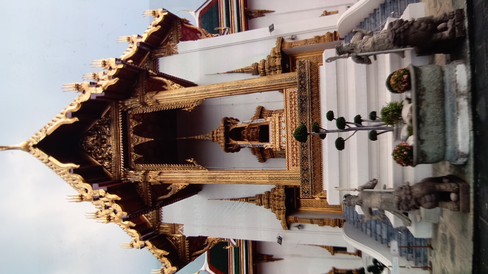 The image shows a section of a grand, ornate temple with intricate gold detailing. The architecture is distinctly Southeast Asian, possibly Thai, given the location context of Koh Phangan, Thailand. The structure features multi-tiered, pointed roofs adorned with gold trim, detailed carvings, and what appear to be decorative finials.
In the foreground, on the right side, are stone statues that resemble guardian figures, placed on either side of a set of white stairs. One statue is visible, holding a staff or spear. In front of the statues are potted plants and decorative bushes, one with small red flowers. The stairs lead up to a white wall, which is also decorated with gold patterns and has arched openings.
The scene suggests a cultural or religious site, likely a temple or palace complex. The lighting indicates daytime, with a bright, overcast sky suggesting it might be early morning or late afternoon, or simply a cloudy day. There is no discernible text in the image. The overall impression is one of grandeur, historical significance, and meticulous craftsmanship. The composition, with the ornate architecture dominating the frame, highlights the beauty and complexity of the building.