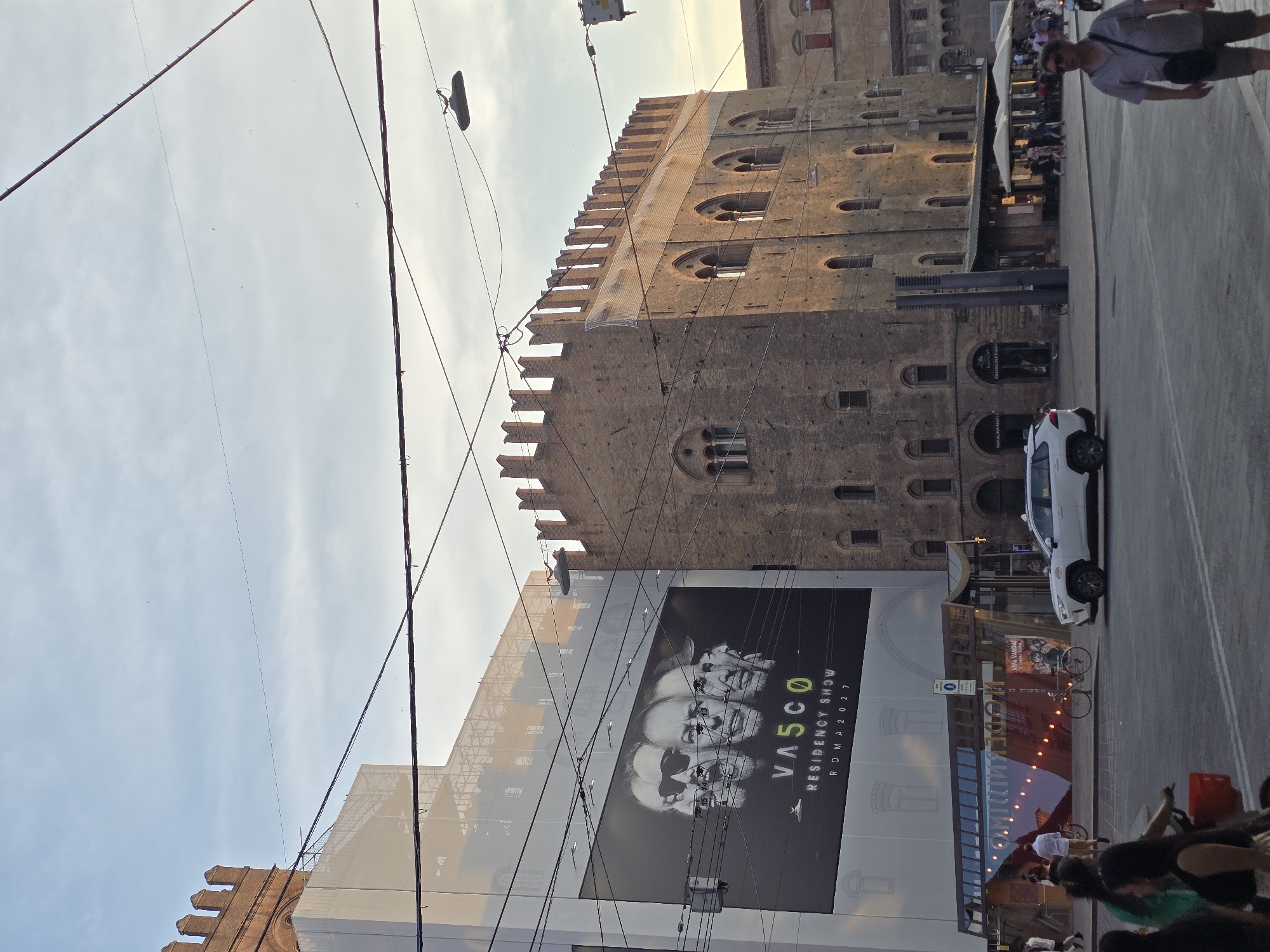 This media file captures a street scene in Bologna, Italy, likely during the late afternoon or early evening as indicated by the warm, low-angled sunlight casting long shadows. The sky is clear with some wispy clouds.
The foreground is dominated by a large billboard featuring a black and white portrait of three men, with the text "VASCO RESIDENCY SHOW ROMA 2027" prominently displayed. This billboard is attached to a modern building, possibly under construction or renovation, indicated by scaffolding and white sheeting. Overhead, a complex network of electrical wires crisscrosses the frame, with some attached to poles and buildings.
Behind the modern building, a historic brick tower, characteristic of Bologna's architecture, rises up. It has crenellated battlements and arched windows. Further in the background, another older building with red accents and arched openings is visible.
On the street, a white car is parked, and a bicycle is leaning against a railing. Several people are visible walking or standing, some in the street and others near outdoor seating areas of a building that appears to be a restaurant or cafe. One man, walking towards the camera, is wearing a grey shirt and shorts, carrying a crossbody bag. The overall atmosphere appears relaxed and casual, typical of a European city street scene. The presence of outdoor dining and pedestrians suggests a vibrant urban environment.