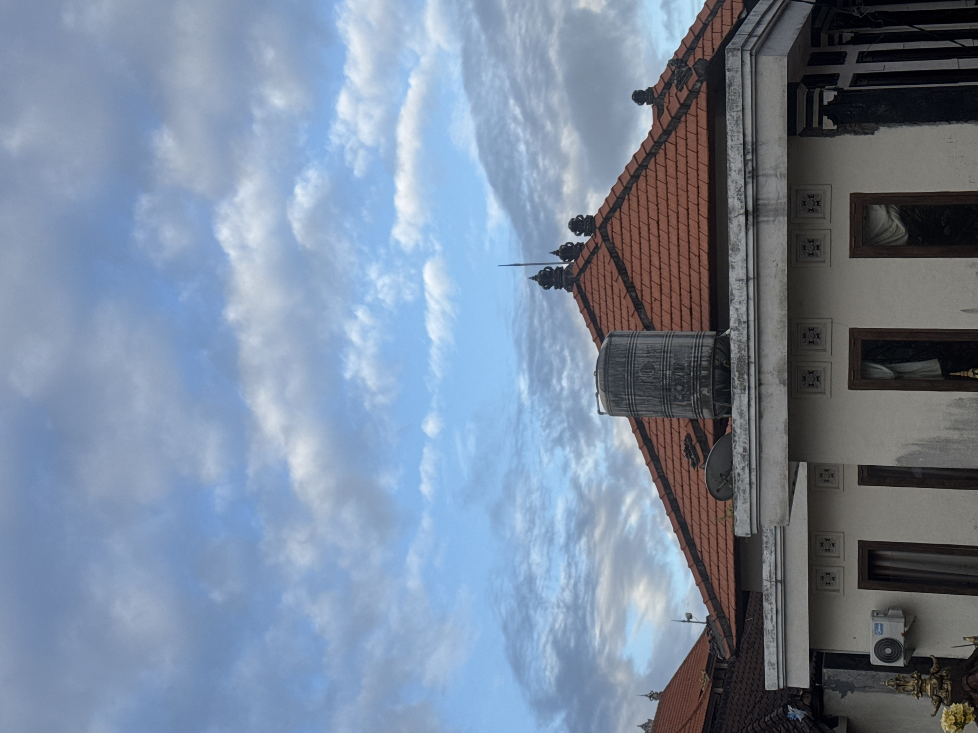The image depicts the rooftops of buildings in Kuta, Indonesia, under a cloudy blue sky. The foreground features a white building with a flat white ledge and an air conditioning unit mounted on its side. Above the white building is a tiled, orange-roofed structure with decorative black elements on the ridge. A large, cylindrical water tank is visible on top of this roof, alongside a satellite dish. Further back, another orange-tiled roof is visible, also with decorative elements. The sky is a mix of blue and gray clouds, with wispy white clouds scattered across.