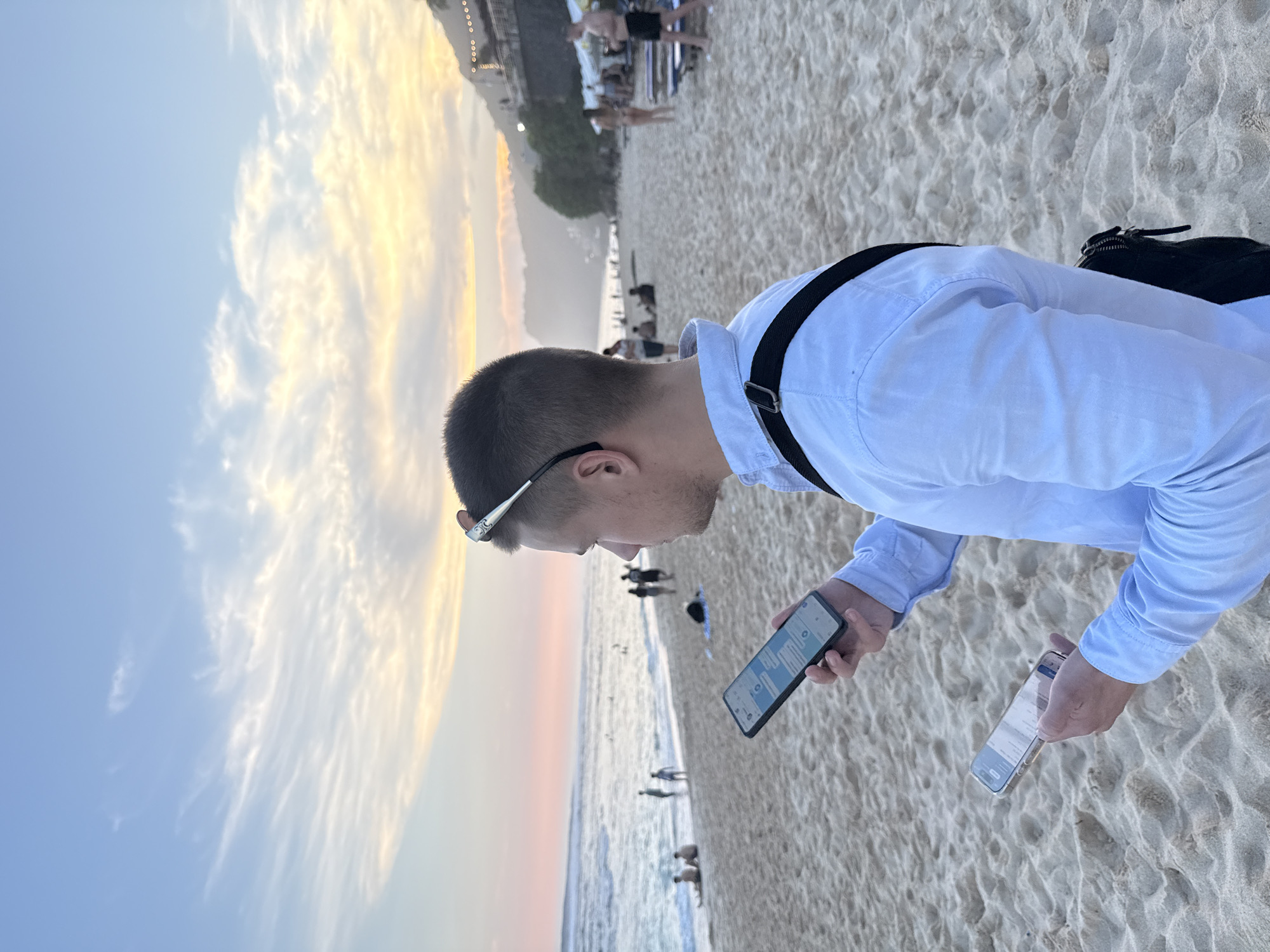 A man is standing on the sand at the beach in Kuta Selatan, Indonesia, holding two phones, with the ocean, beachgoers, and a setting sun in the background.