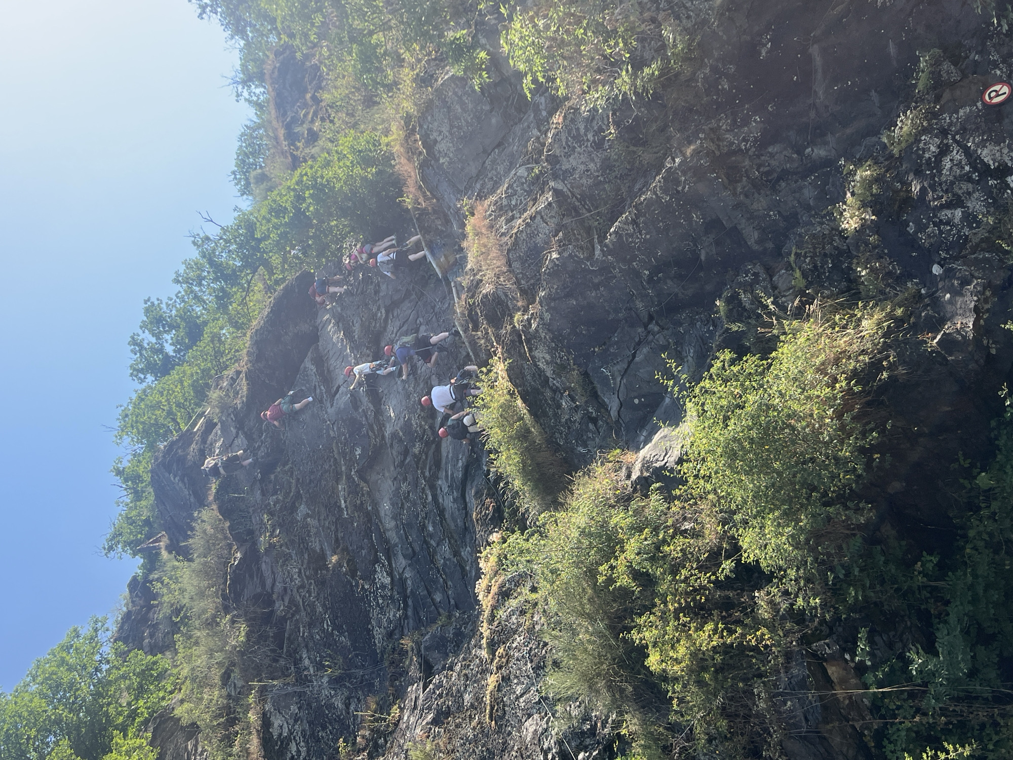 A group of people in red helmets are climbing a steep, rocky cliff face under a clear blue sky. The cliff is covered in dark gray rock and patches of green vegetation. Several individuals are visible at different points on the cliff, appearing to be engaged in a guided climbing activity. Some are further up the cliff, while others are closer to the bottom. The scene suggests an outdoor recreational activity taking place in a natural environment.